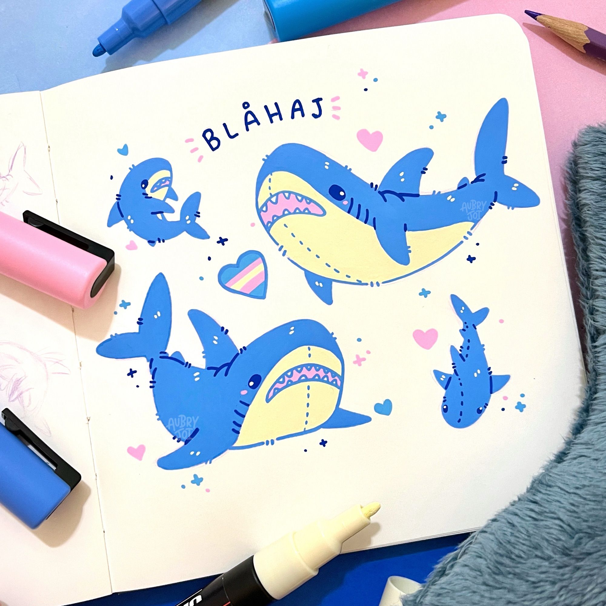 A page of a sketchbook showing several drawings of the plush shark “Blahaj,” aka the IKEA shark. The doodles are all drawn with paint pens and show bright, solid colors. There’s a small heart with trans flag stripes towards the center of the page, and at the top is the word “Blahaj.”