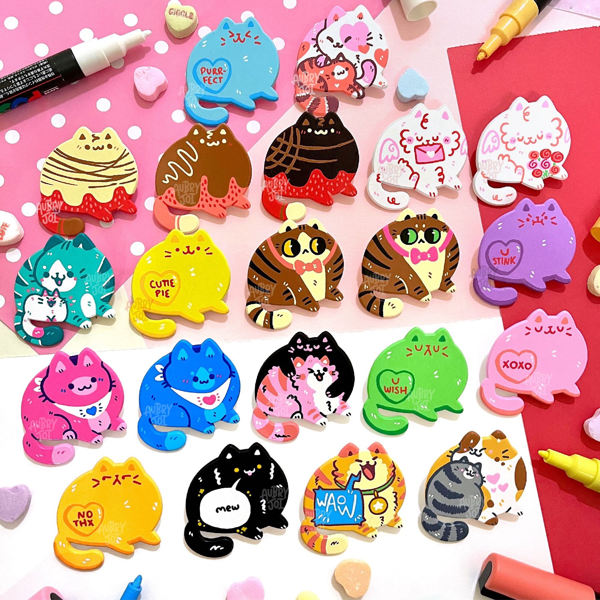 A photo of 21 colorful, hand-painted fat cat enamel pins. Each one is unique. Most are Valentine’s Day themed, in colors like pink, white, red, & brown. Some look like chocolate covered strawberries; others look like conversation heart candies; some are just colorful. There are Posca markers & candy hearts around the pins.