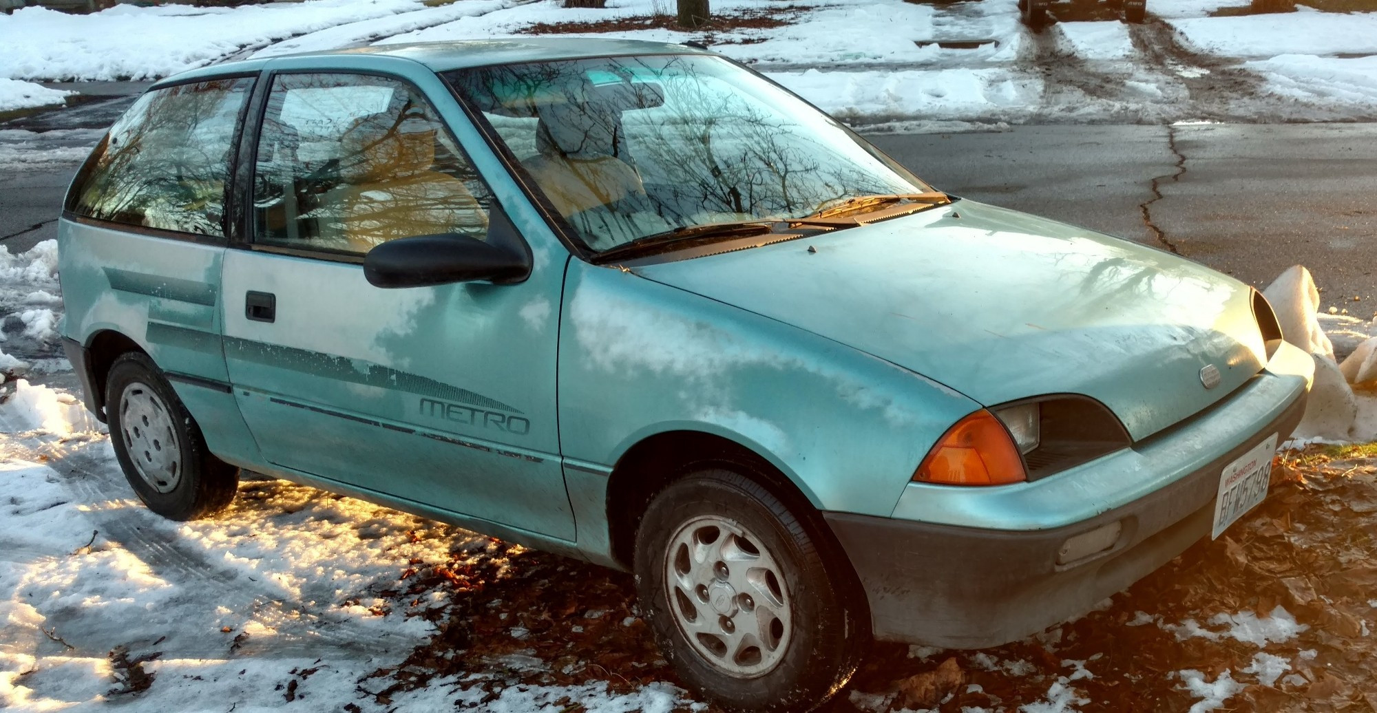 A 1991 teal Geo Metro parked on the sidewalk, crooked. There is snow on the ground and it is golden hour