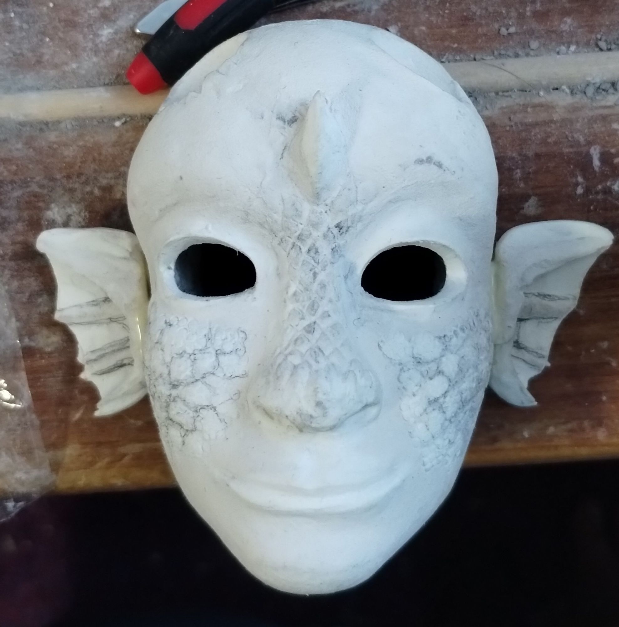 A humanoid doll with scales across the bridge of the nose forehead and cheekbones. There are now fin like ears glued to the sides of the head