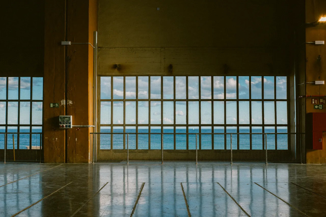 A large window inside an empty industrial building, offering a view of the ocean through the grid-like panes.