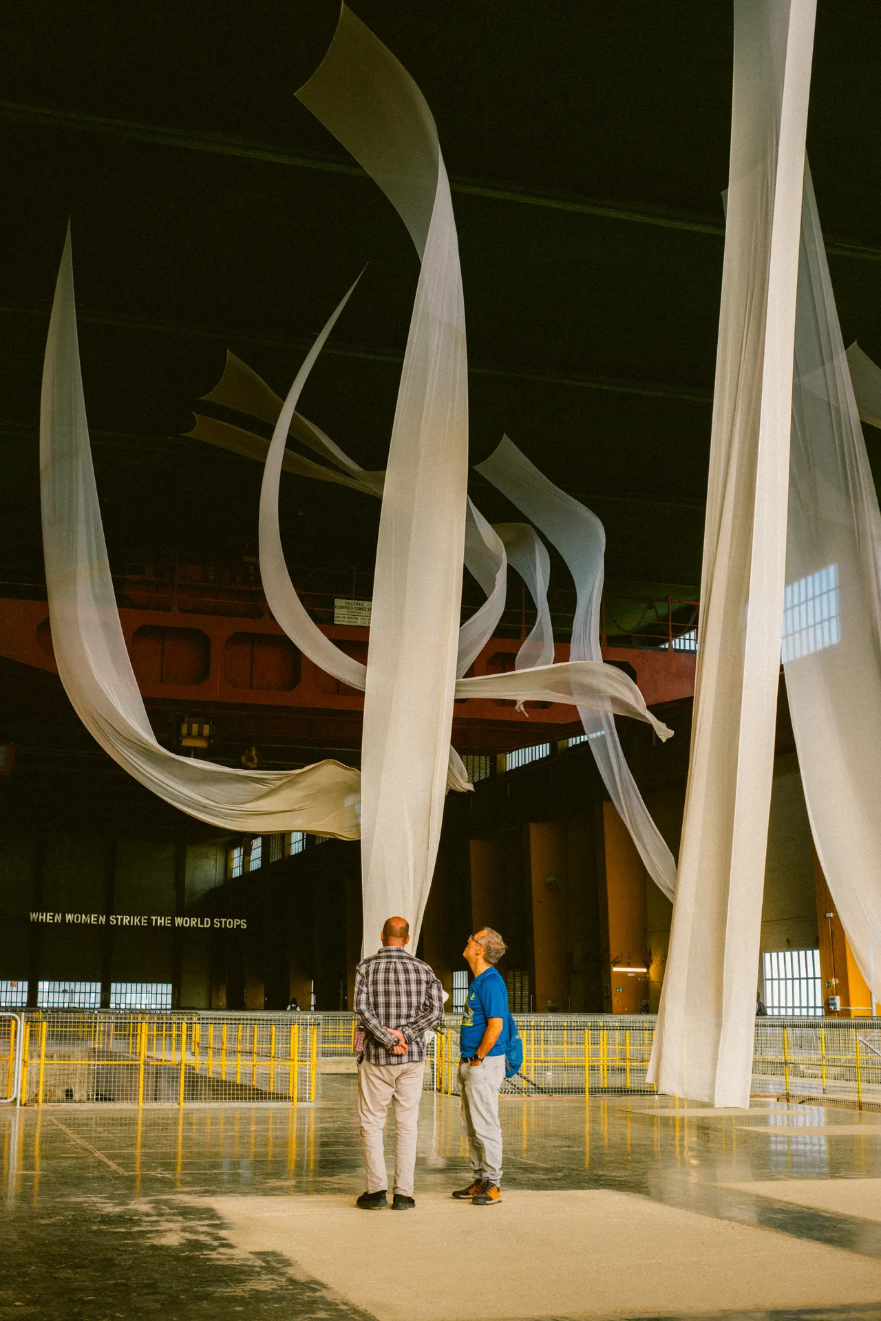 Two men observe flowing white fabric suspended from the ceiling in a large industrial space, with a banner in the background that reads "When women strike, the world stops."