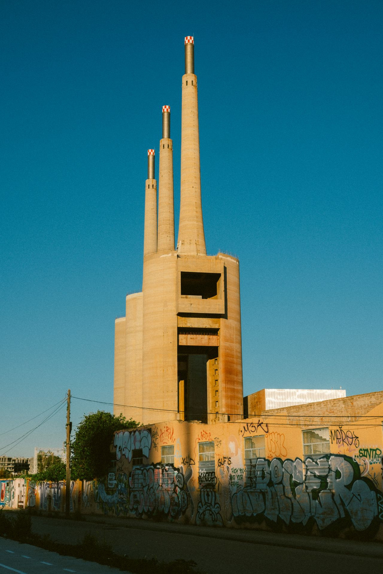 A tall, industrial building with three cylindrical smokestacks, surrounded by graffiti-covered walls under a clear blue sky.