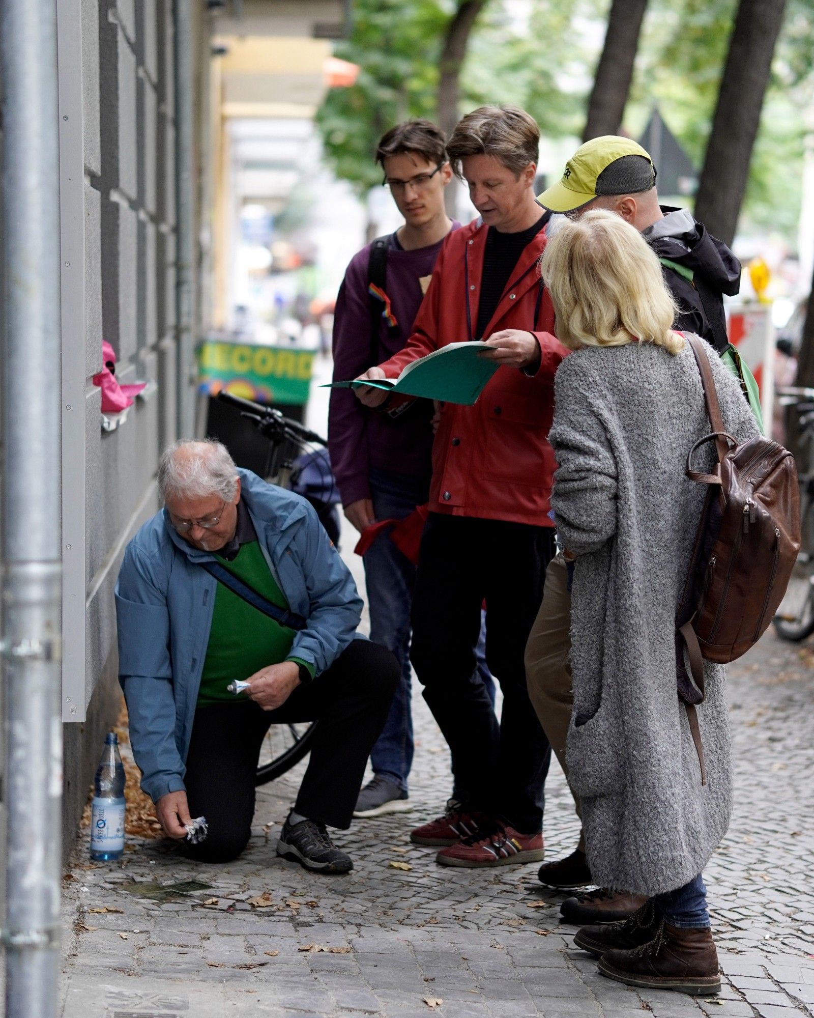 Fünf Personen auf einem Bürgersteig. Sie stehen um drei in den Bürgersteig eingelassene Stolpersteine herum. Eine Personen kniet und hält Putztücher in der Hand, eine andere liest aus einem Hefter vor.