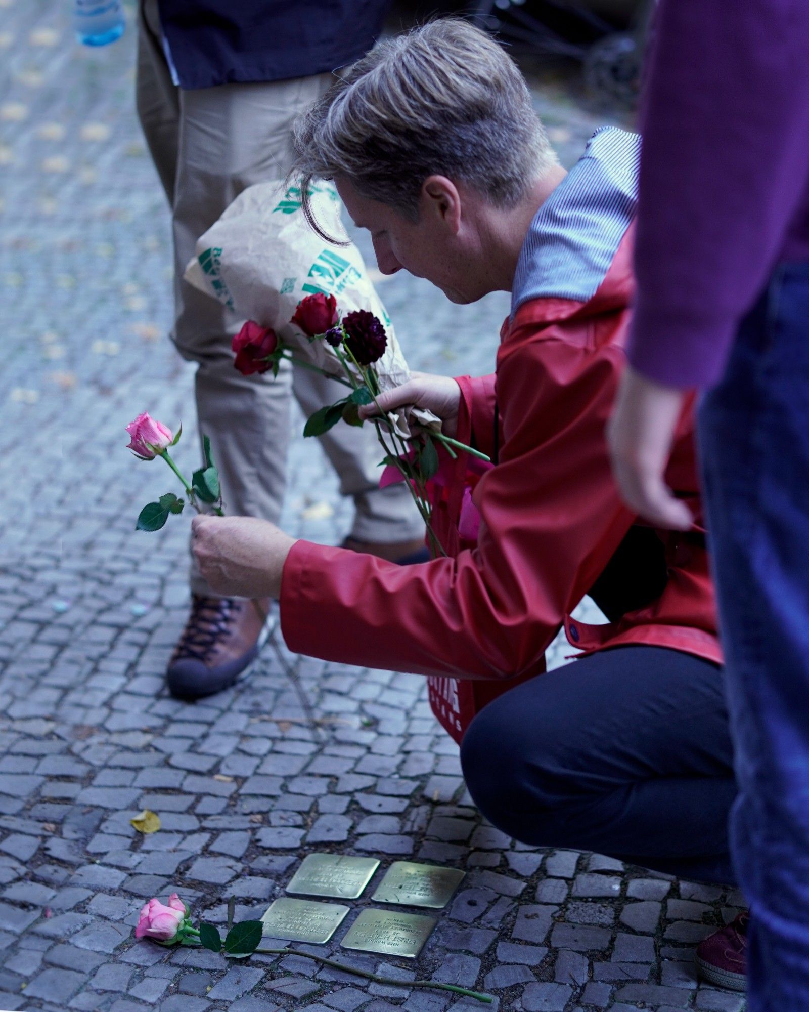 Eine Person, die auf einem Bürgersteig hockt und eine Blumen in der Hand hält. Auf dem Boden liegt eine Rose neben vier in den Bürgersteig eingelassenen Stolpersteinen.