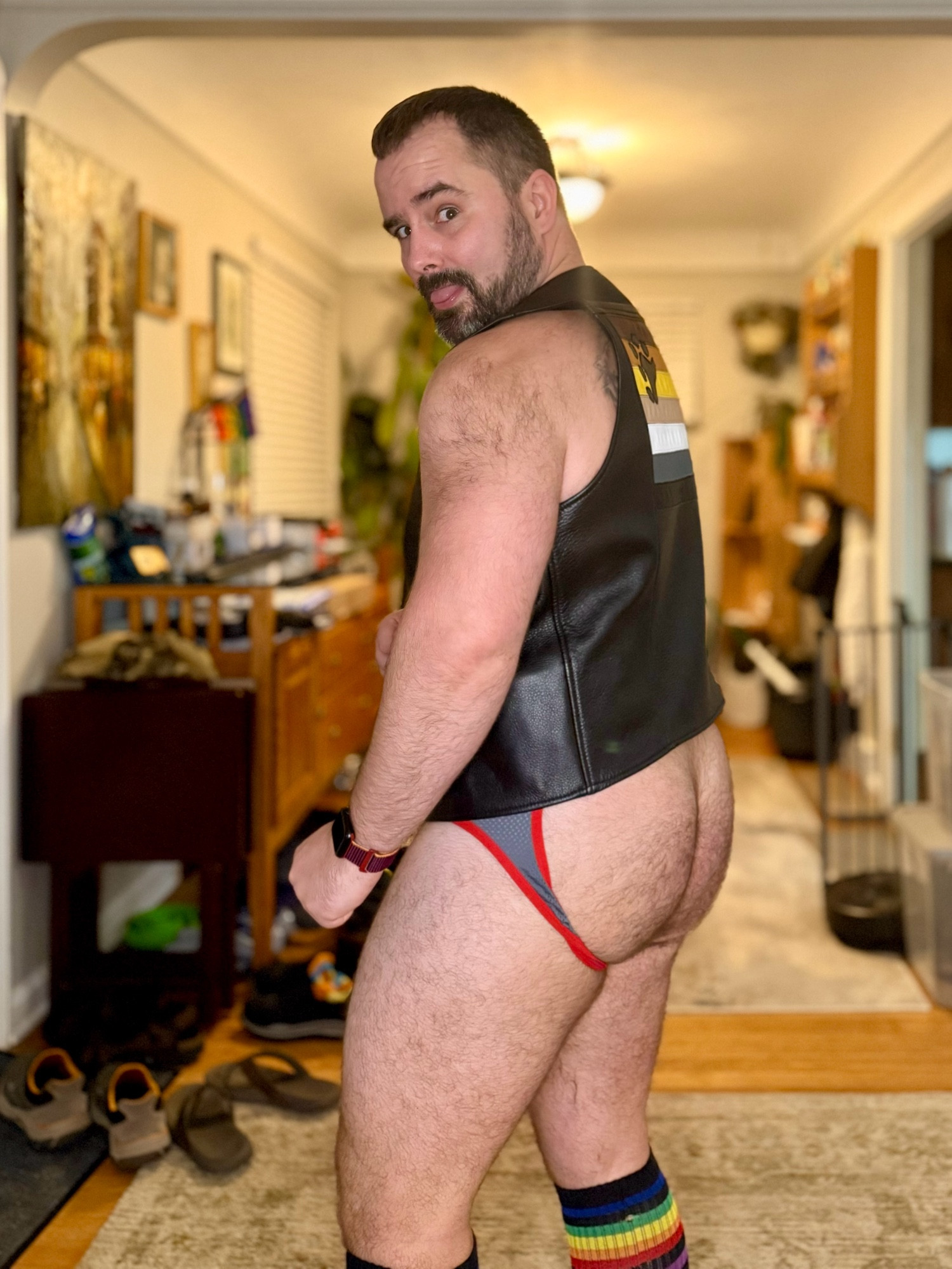 Photo of me in a mesh jockstrap and a black leather vest. The back of the vest has a bear pride flag design in leather.  My hairy butt is visible.