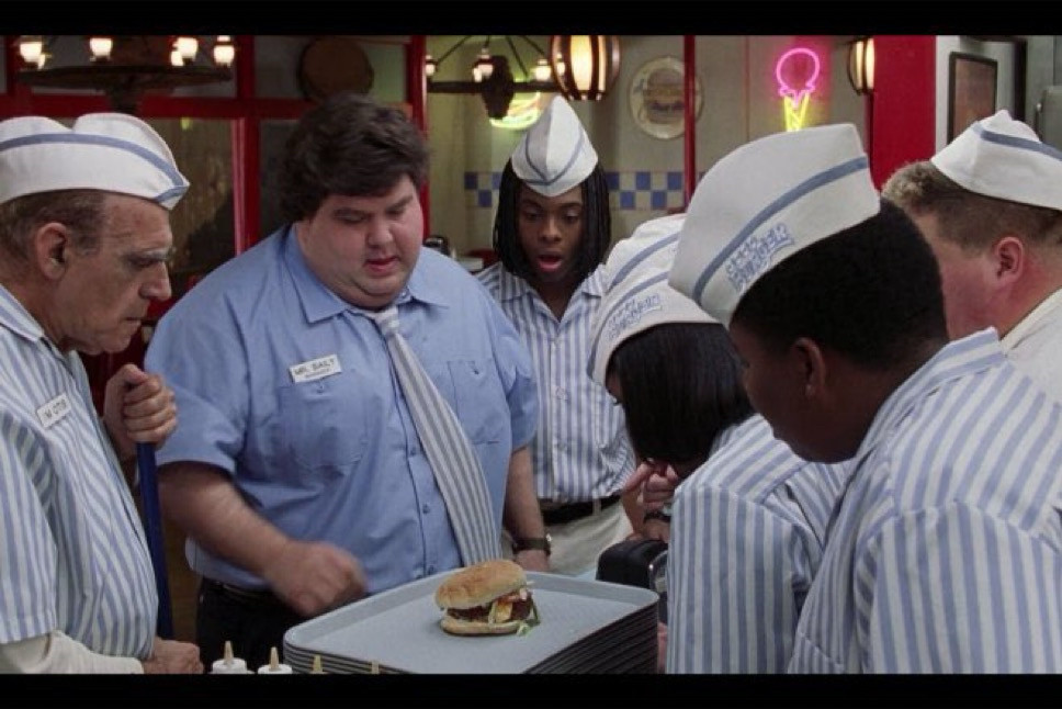 Scene from Good Burger (1997), in which a group of fast food burger restaurant employees huddle around a burger from a competitor’s restaurant with looks of shock and concern on their faces.