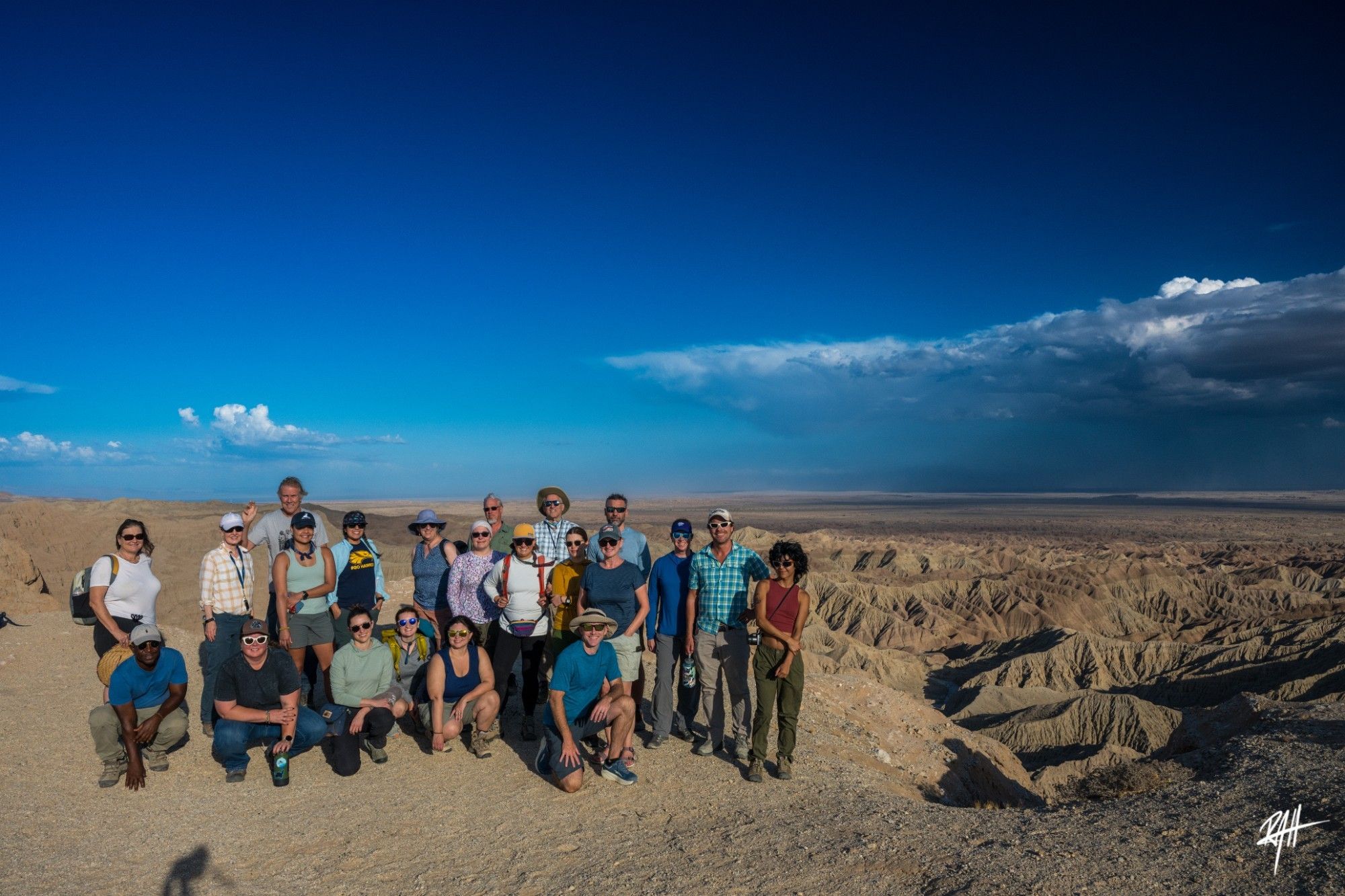 Pre-GSA field trip group in front of the Fonts Point lookout. 22 people squint into the sun, with dramatic badlands and a storm behind them.