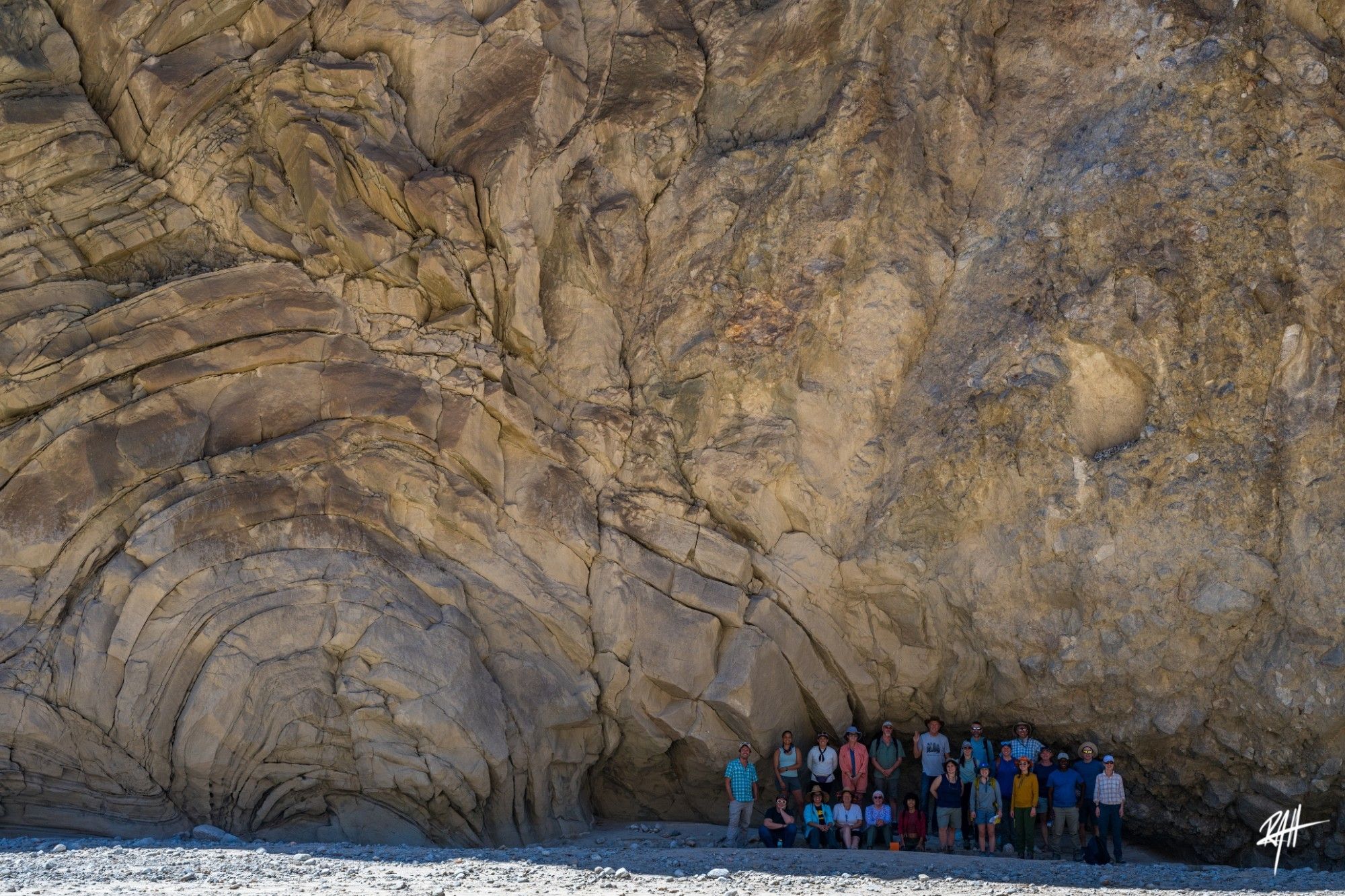 Pre-GSA field trip group in front of the sturzstrom/squooshed turbidite contact, Fish Canyon Wash. A group of 22 people stand in front of a cliff. On the right of the cliff is a mass of angular granite fragments. On the left are tan sandstones and shales bent into anticlinal orientations.