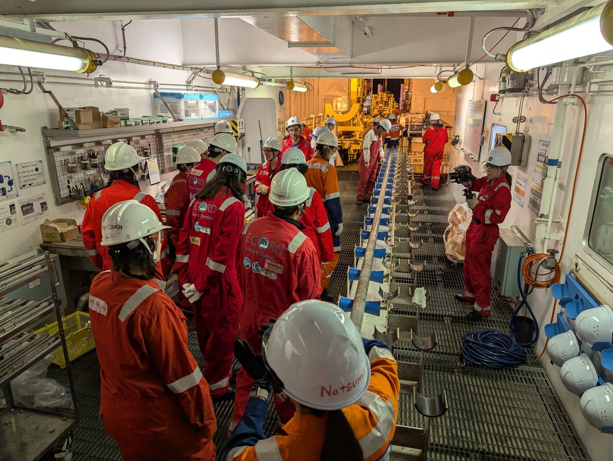 A bunch of scientists in full PPE gather around the core, which is laid out on a core-holding device in an industrial lab space.