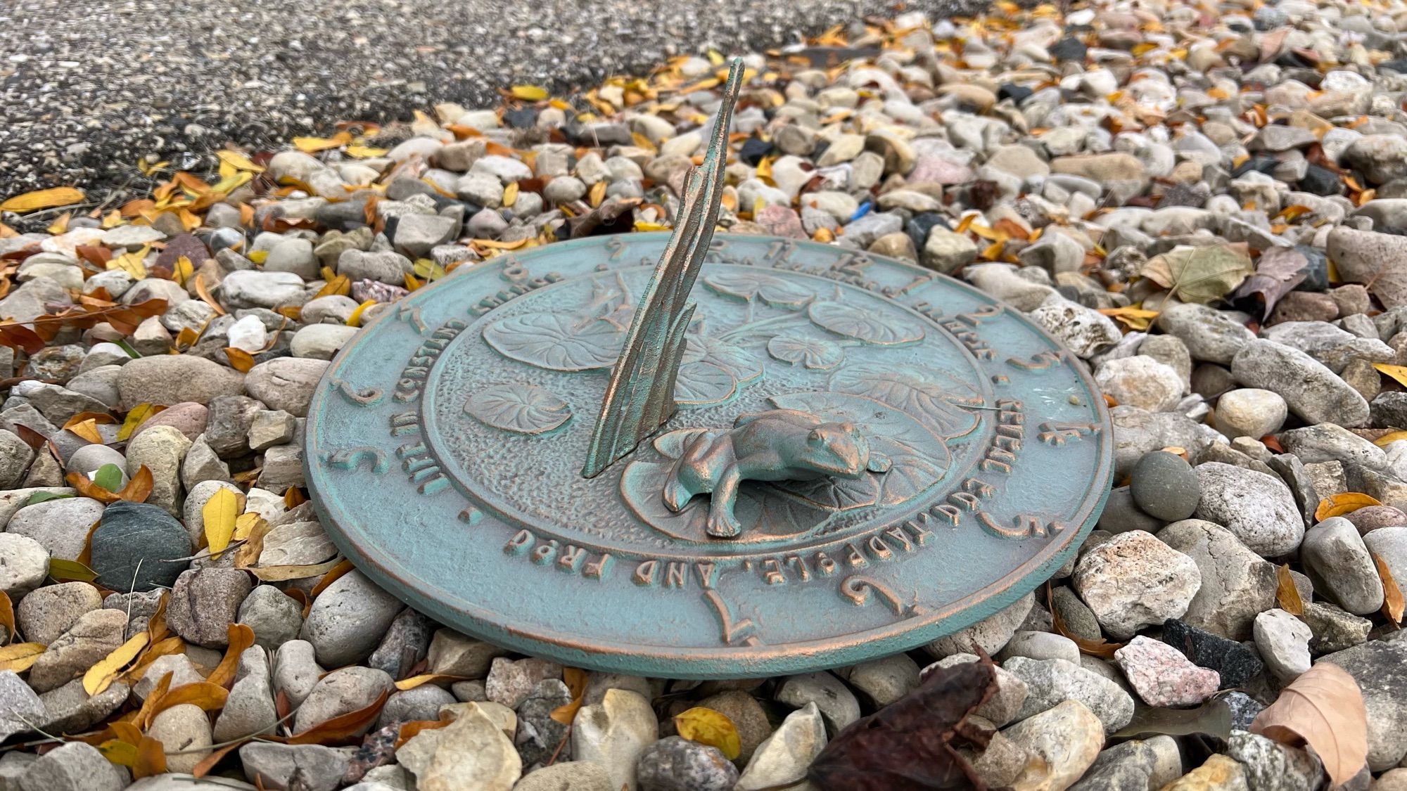 a metal sundial with a frog ornament atop some rocks