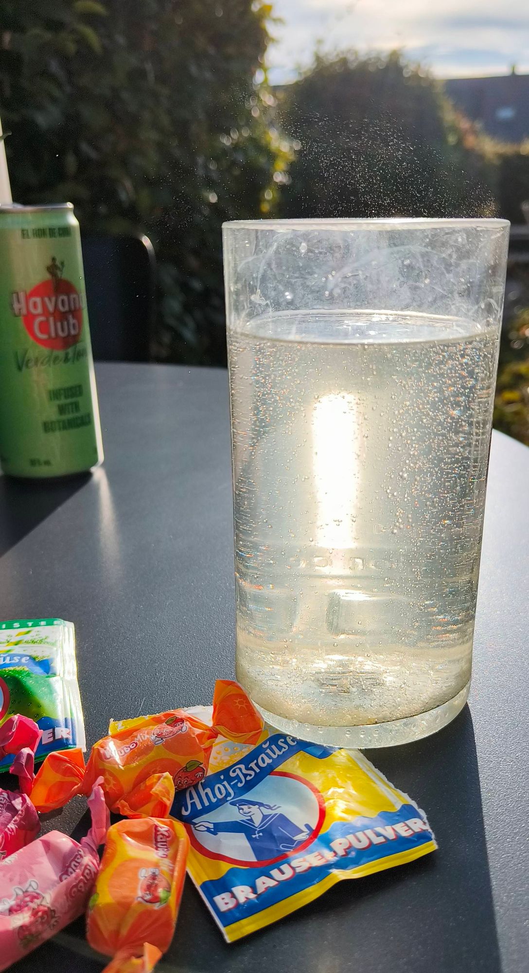 Auf einem Tisch draußen in der späten Nachmittagssonne: Ein Glas mit perlendem, nahezu klaren Getränk durch das die Sonne scheint, davor Bonbons und Brausepulver. Im Hintergrund eine Dose Rum-Tonic.