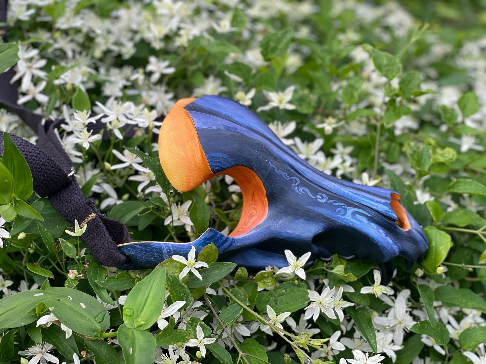 A painted canine skull mask rests among small white star flowers. The mask in painted in blues and yellows in the eye sockets.