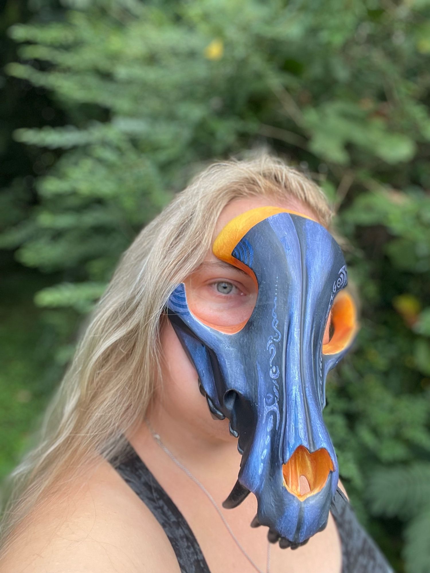 A painted canine skull mask being worn. The mask in painted in blues and yellows in the eye sockets. The wearer is looking into the camera. Ash blonde hair cascades around the face.