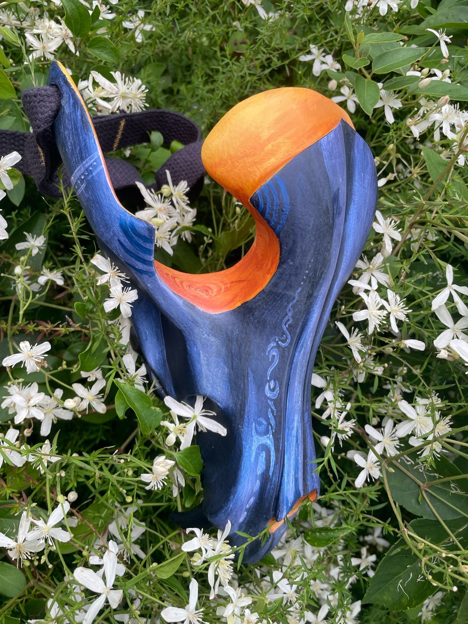 A painted canine skull mask rests among small white star flowers. The mask in painted in blues and yellows in the eye sockets.