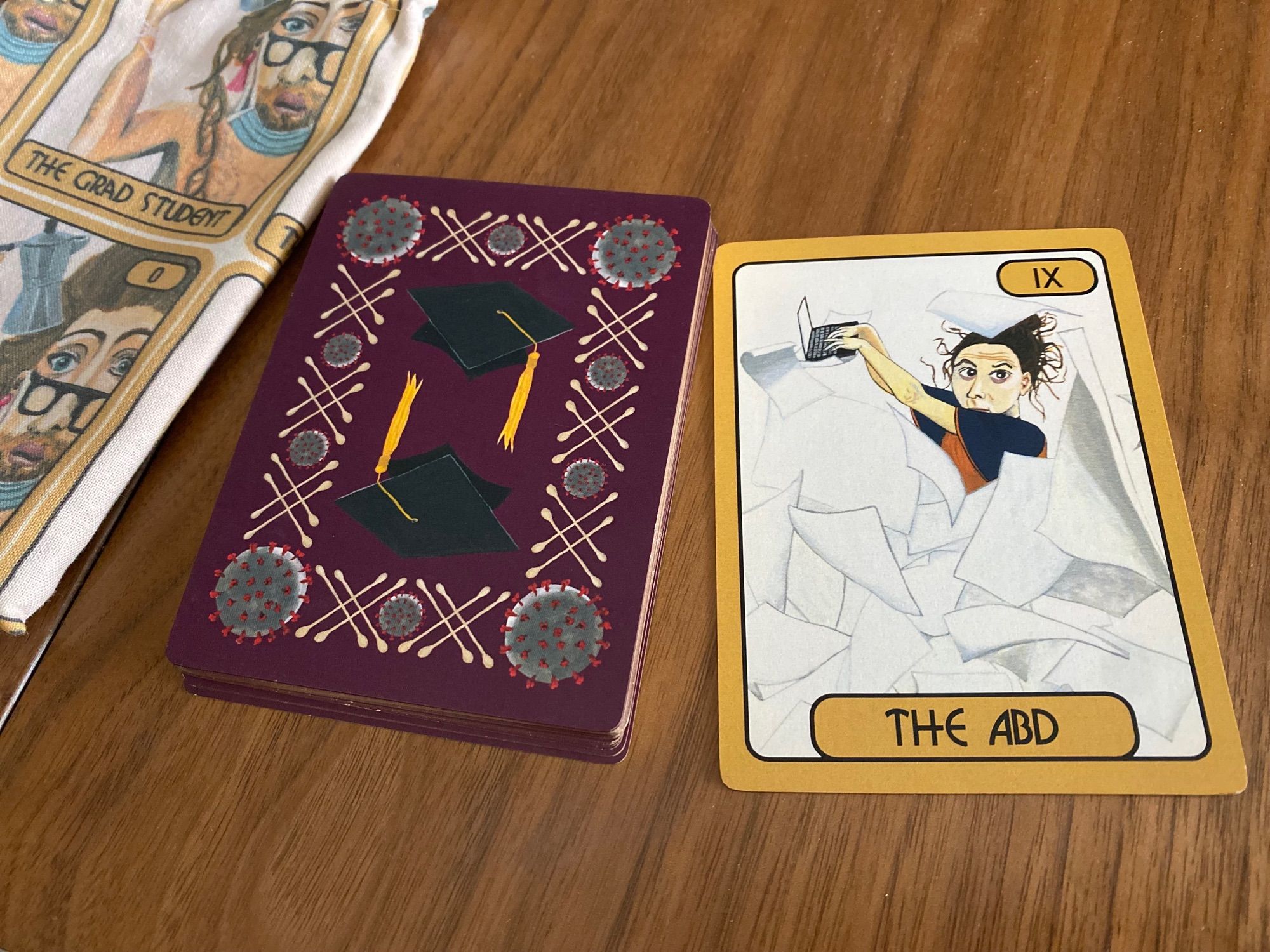 A deck of cards lies on a wood table, the stack of cards is turned down, and the back of the cards have artwork with the following imagery: a graduate cap, a virus, and Covid-19 test swabs. One card is turned over, and the label at the bottom of this card says “The ABD”. The artwork on the card is abstract; a human figure is mostly obscured by a cascade of paper, and they face towards us with kind eyes, messy hair, and hands that are typing on a laptop.