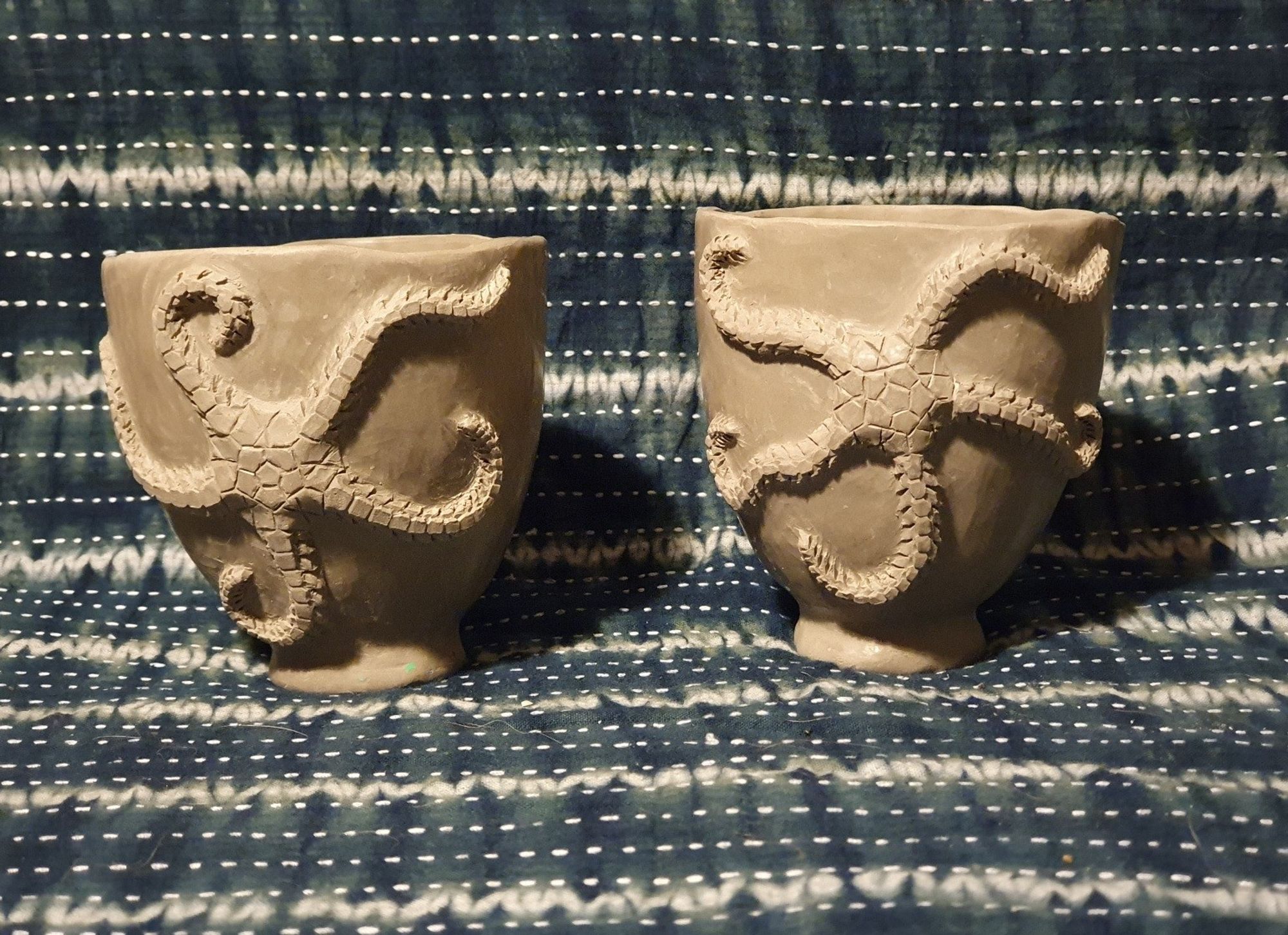 Two greenware pottery cups with sculpted relief brittlestars on a striped fabric back. The brittlestars have undulated arms and are almost as large as the cups