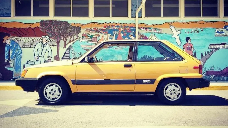 A yellow Toyota tercel with a graffiti backdrop