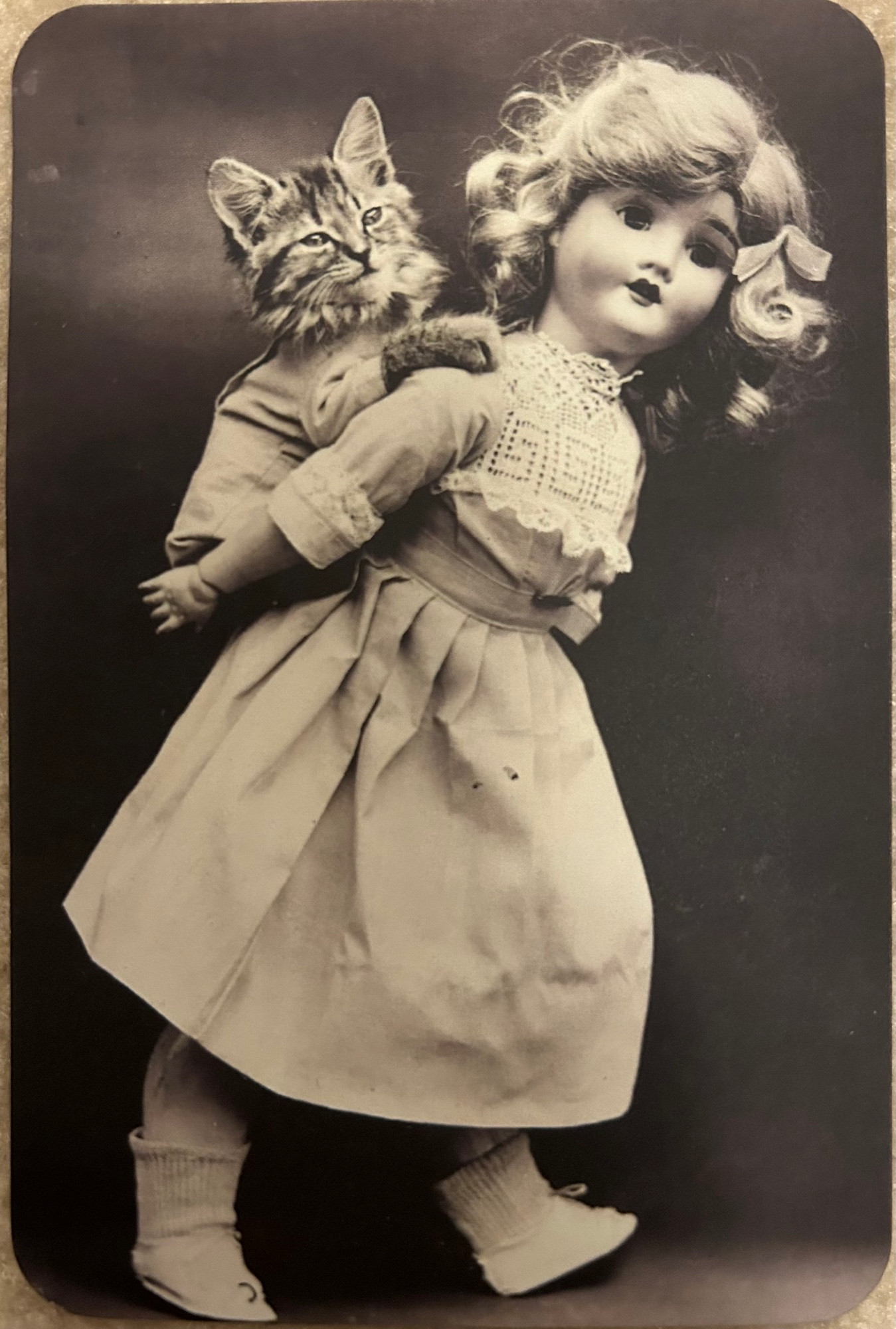 Black and white photo of an antique doll with blonde curls and a pale dress giving a piggy back ride to a smug tabby kitten wearing a little jacket