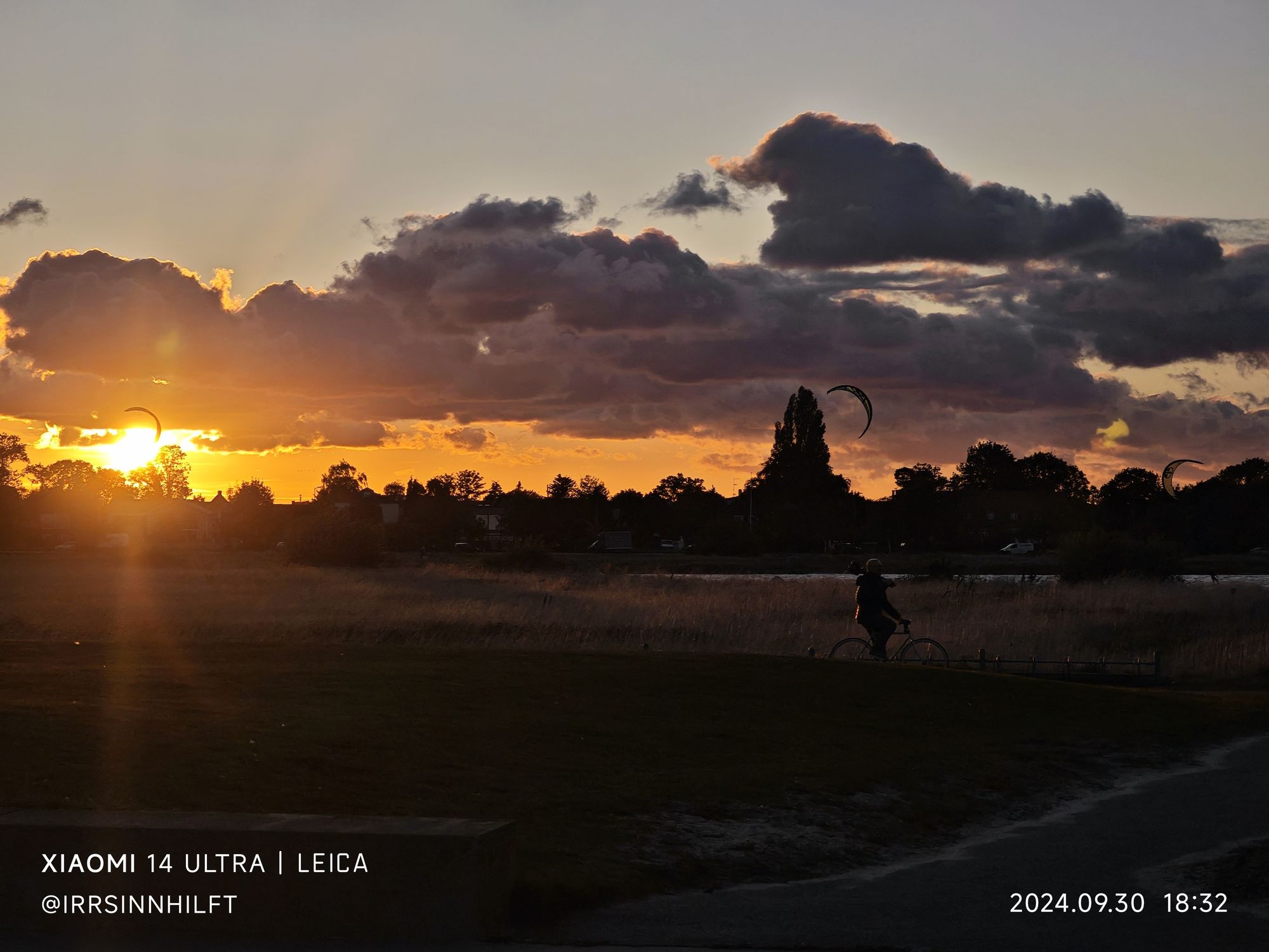 Zwischen Wolken und bewaldetem Horizont scheint die Abendsonne auf das Dünengras im Park. Die Schatten von 3 Kitesurf-Segeln sind zu sehen und eine Person auf dem Fahrrad verlässt das Bild nach rechts