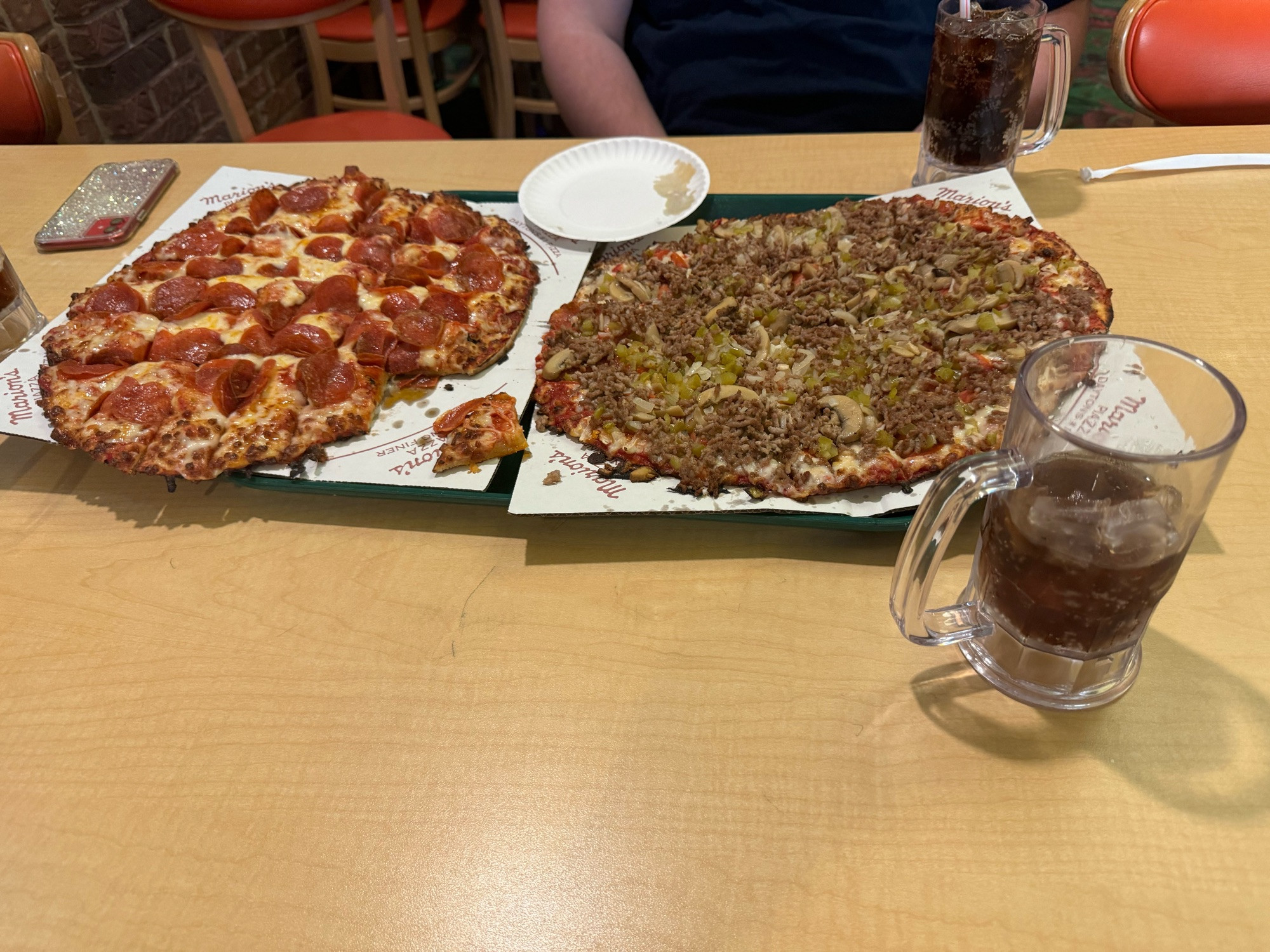 A tray with two thin-crust pizzas from Marion’s Piazza, one is pepperoni and the other is supreme, covered in a layer of sausage. There’s also a mug of rootbeer next to the trays