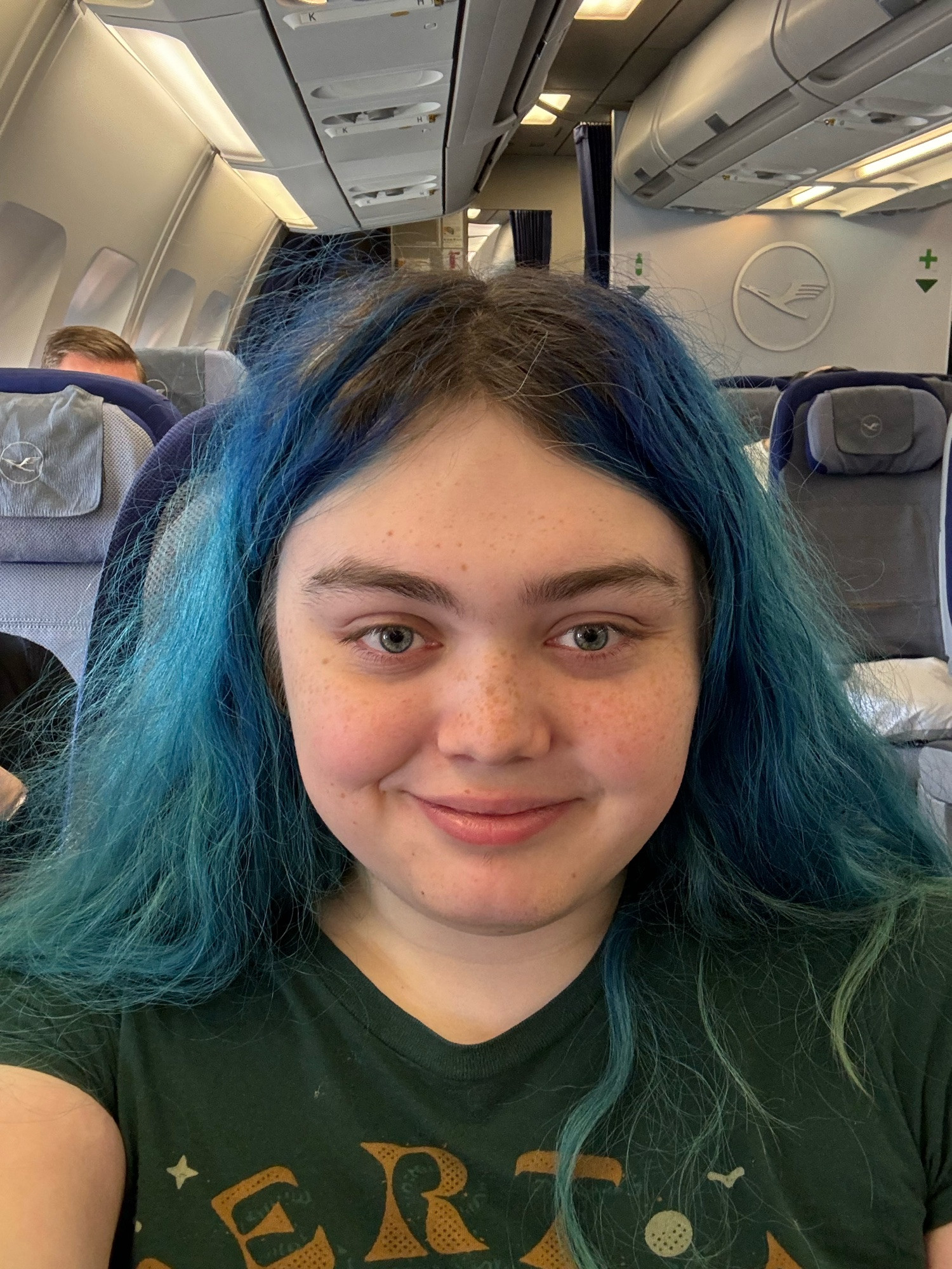 A selfie of me sitting in an Airbus A340-300, on the aisle seat of the right side. There is a Lufthansa logo in white on the wall behind me.