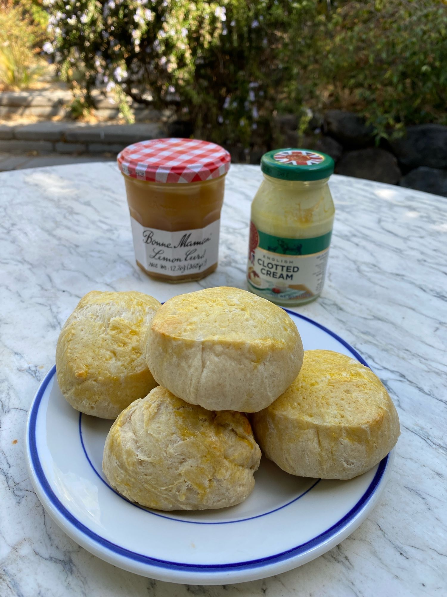 A plate piled with golden brown biscuits o. A marble table with a jar of clotted cream & lemon curd. A blossoming bush with lavender purple flowers in the background.