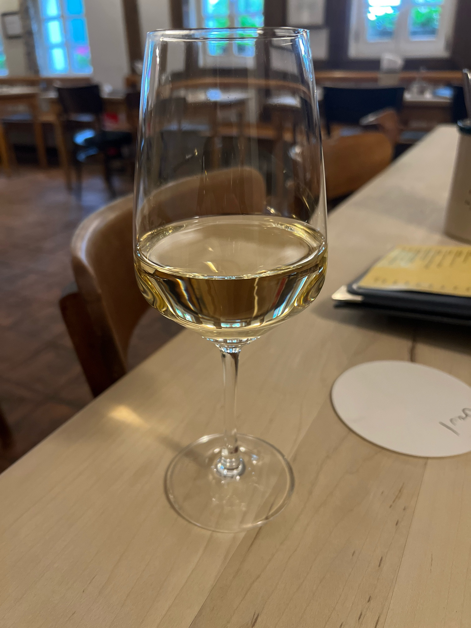 A glass of white wine on a wooden table, chairs and things in the background. It’s a nice restaurant.