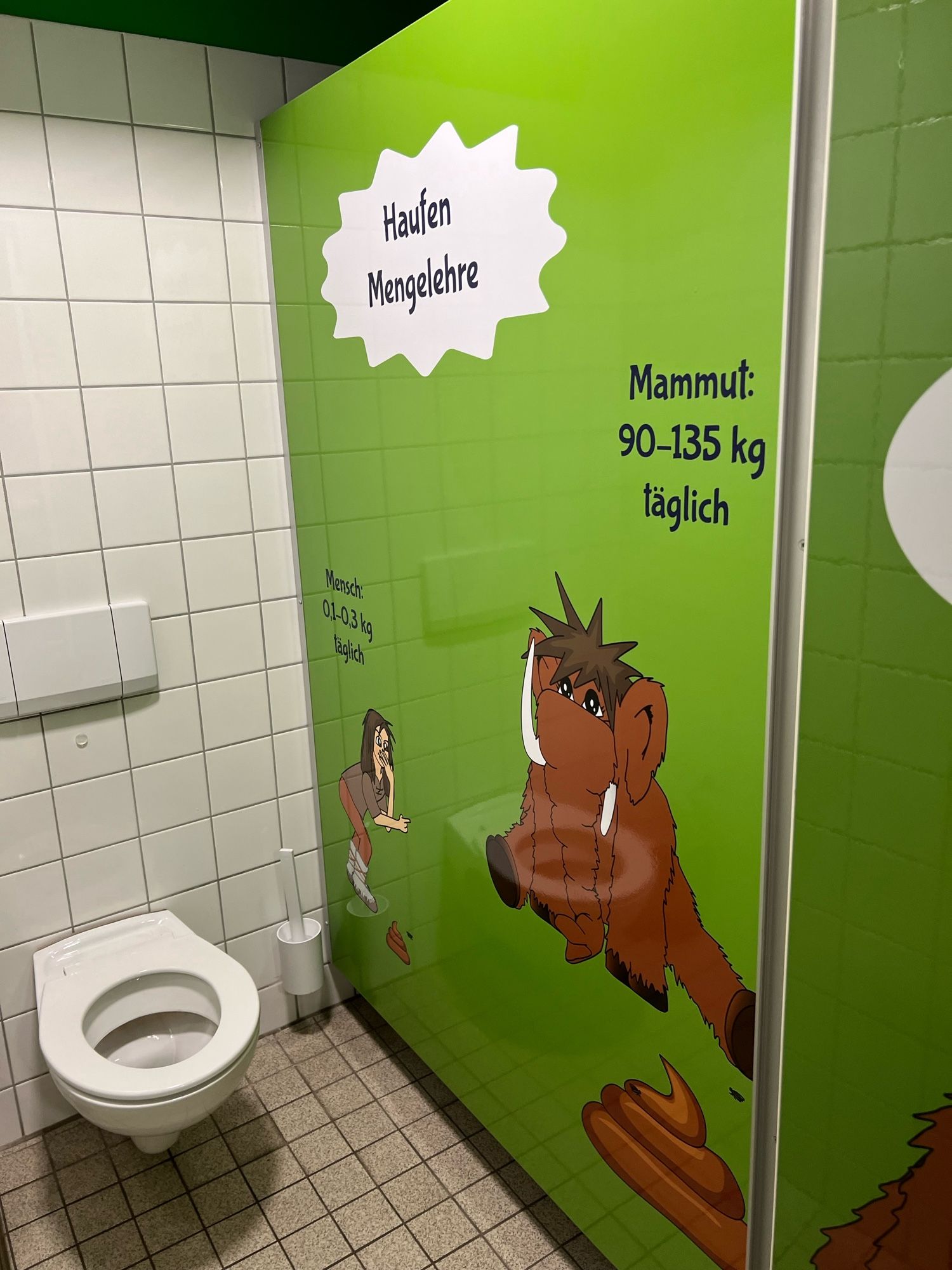 View into a toilet stall with a drawing of a human and a mammoth on the right, with the information that mammoths poop 90-135kg per day.