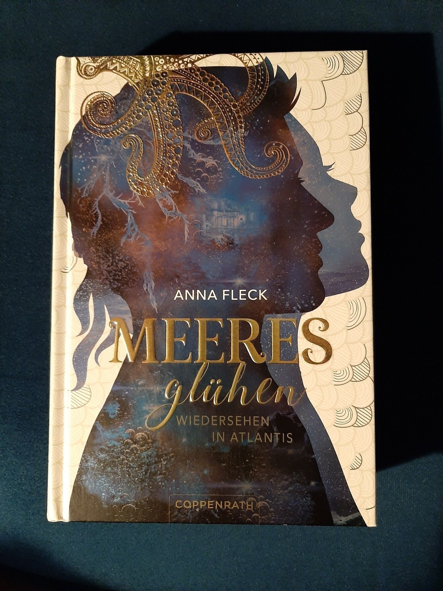 Das Bild zeigt das Buchcover des Romans "Meeresglühen - Wiedersehen in Atlantis" von Anna Fleck. Das Design des Covers ist überwiegend in Blautönen gehalten. Im Vordergrund sieht man das seitliche Profil einer weiblichen und einer männlichen Silhouette, die den Himmel und das Meer in sich aufnehmen. Ein großer, goldener Oktopus ist im oberen Teil des Covers abgebildet und breitet seine Arme über den Titel aus. Im Hintergrund sind die Umrisse einer antiken Stadt zu erkennen. Der Titel ist in goldenen Lettern geschrieben, während der Untertitel "Wiedersehen in Atlantis" in kleineren, schimmernden Buchstaben darunter steht.