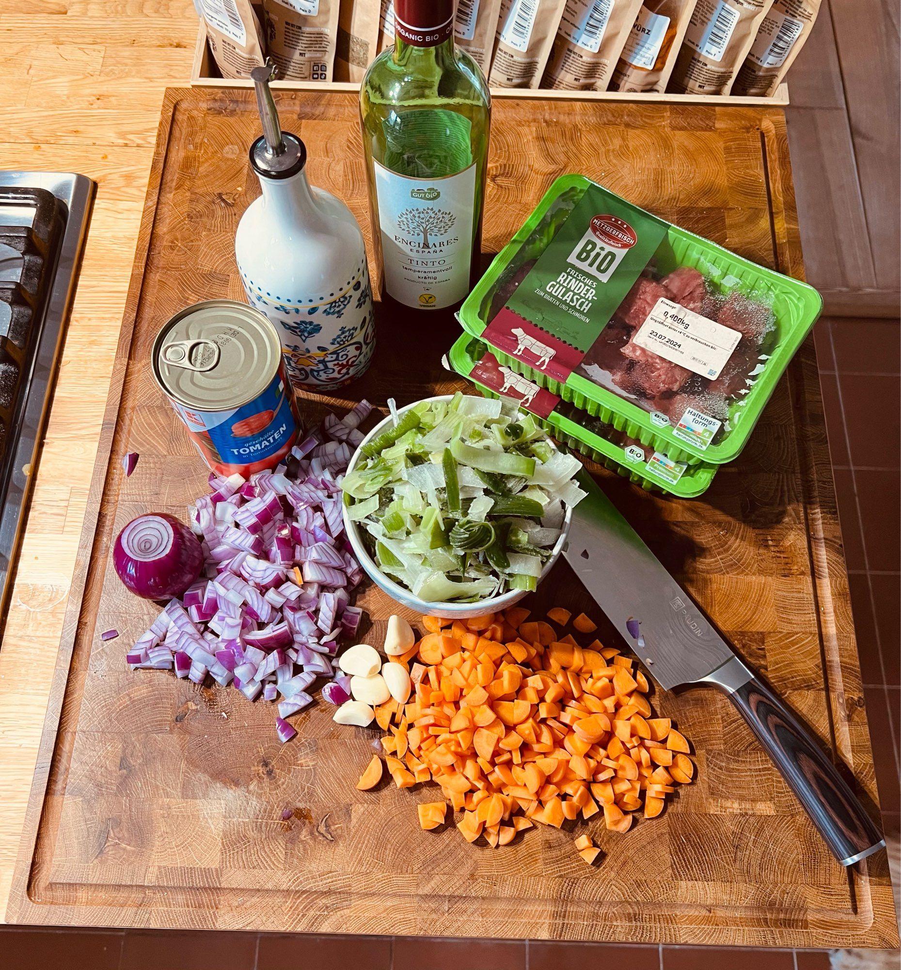 Auf dem Foto ist ein Schneidebrett zu sehen, auf dem verschiedene Zutaten und Küchenutensilien für die Zubereitung einer Mahlzeit liegen. Darauf befinden sich:

- Gehackte rote Zwiebeln
- Geschnittene Karotten
- Mehrere Knoblauchzehen
- Ein Becher mit gehacktem Lauch
- Eine Dose Tomaten (passierte oder gehackte Tomaten)
- Eine Flasche Olivenöl
- Eine Flasche Rotwein (Bio)
- Eine Packung Bio-Rindergulasch
- Ein großes Messer zum Schneiden