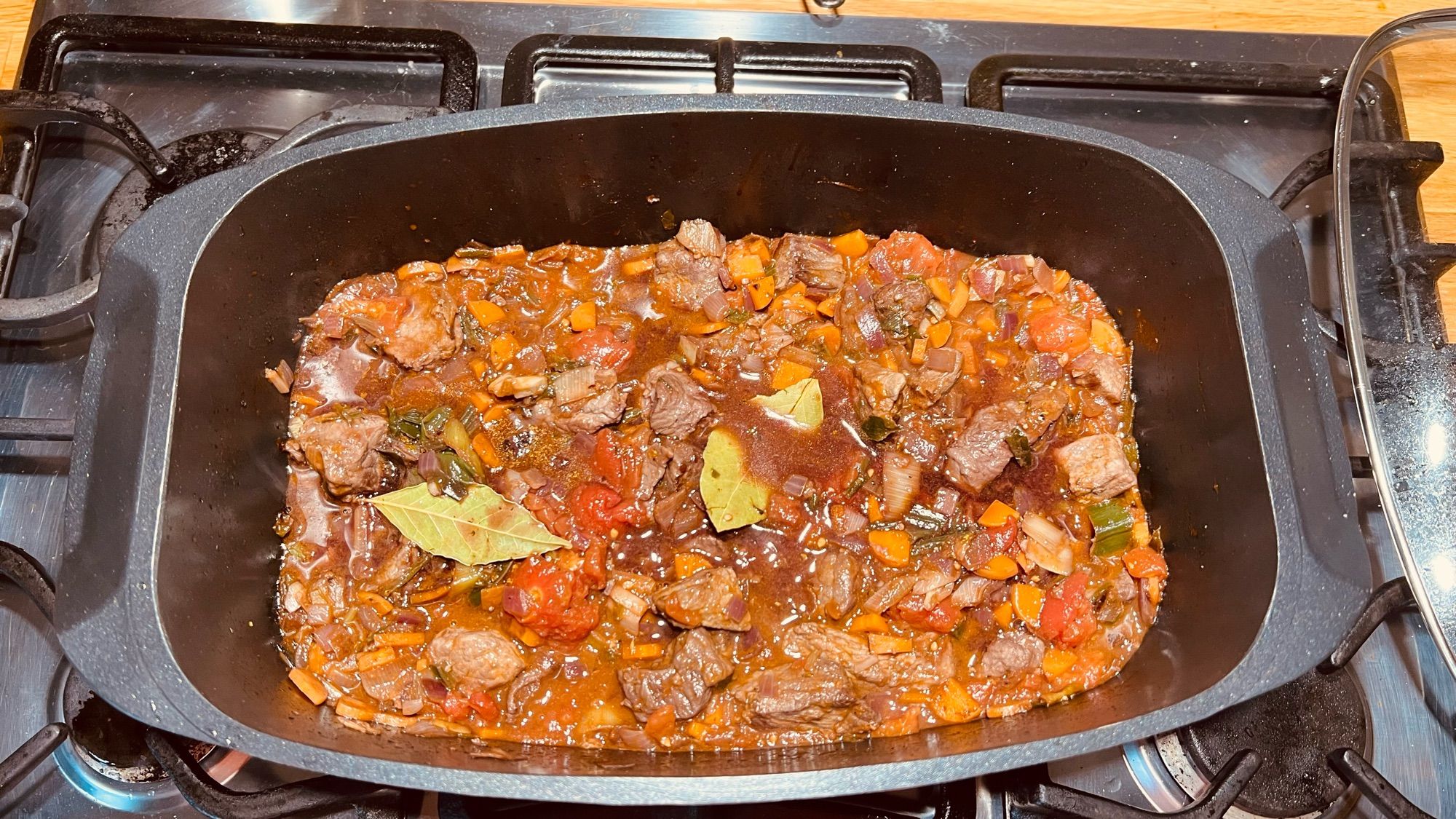 Das Bild zeigt einen Bräter auf einem Gasherd, in dem ein herzhaftes Schmorgericht zubereitet wird. Zu sehen sind Stücke von Rindfleisch, die in einer reichhaltigen, roten Sauce gekocht werden, die aus Tomaten, Karotten, Zwiebeln und wahrscheinlich Sellerie besteht. Zudem sind Lorbeerblätter und andere Kräuter als Gewürze hinzugefügt. Es scheint sich um ein traditionelles Schmorgericht wie Gulasch oder einen Rindfleischeintopf zu handeln, der langsam auf dem Herd gekocht wird.