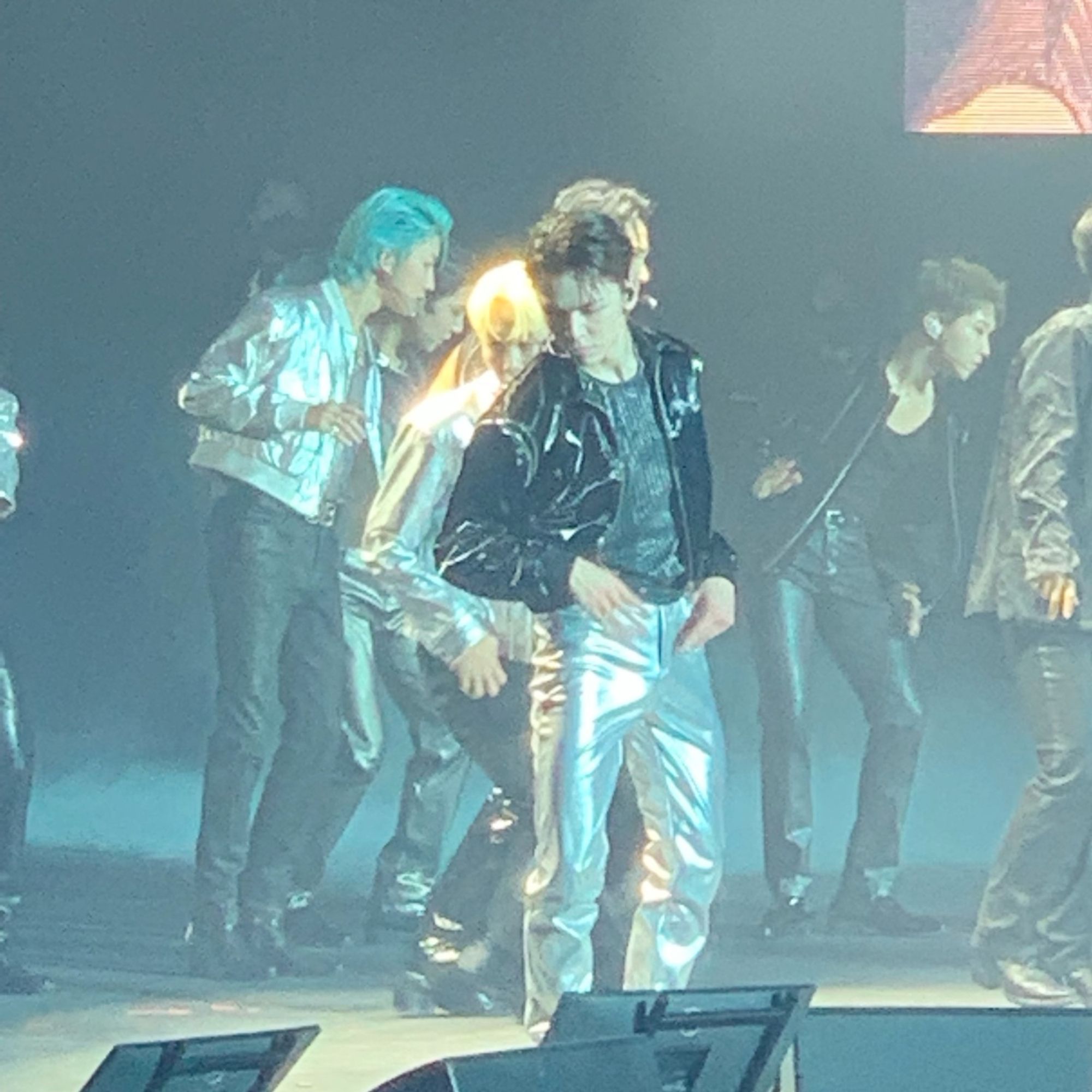 Photo of a person on stage dressed in a shiny black jacket and shiny silver pants with their hands on their hips and head turned to face down. Other people in similar themed outfits in the background are in similar poses facing the opposite direction.