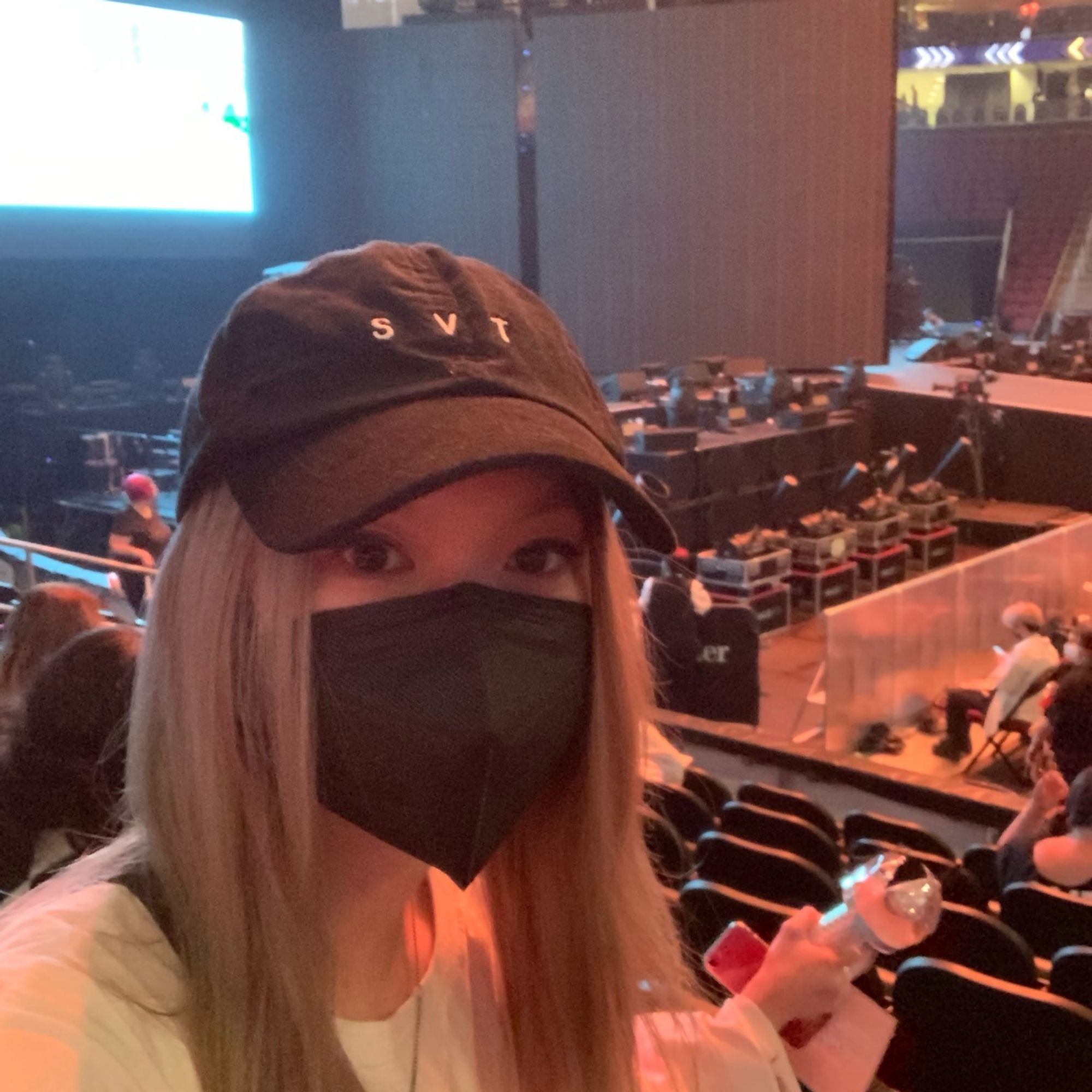 Photo of a blonde person with a black face mask and black cap at a concert stage in the audience