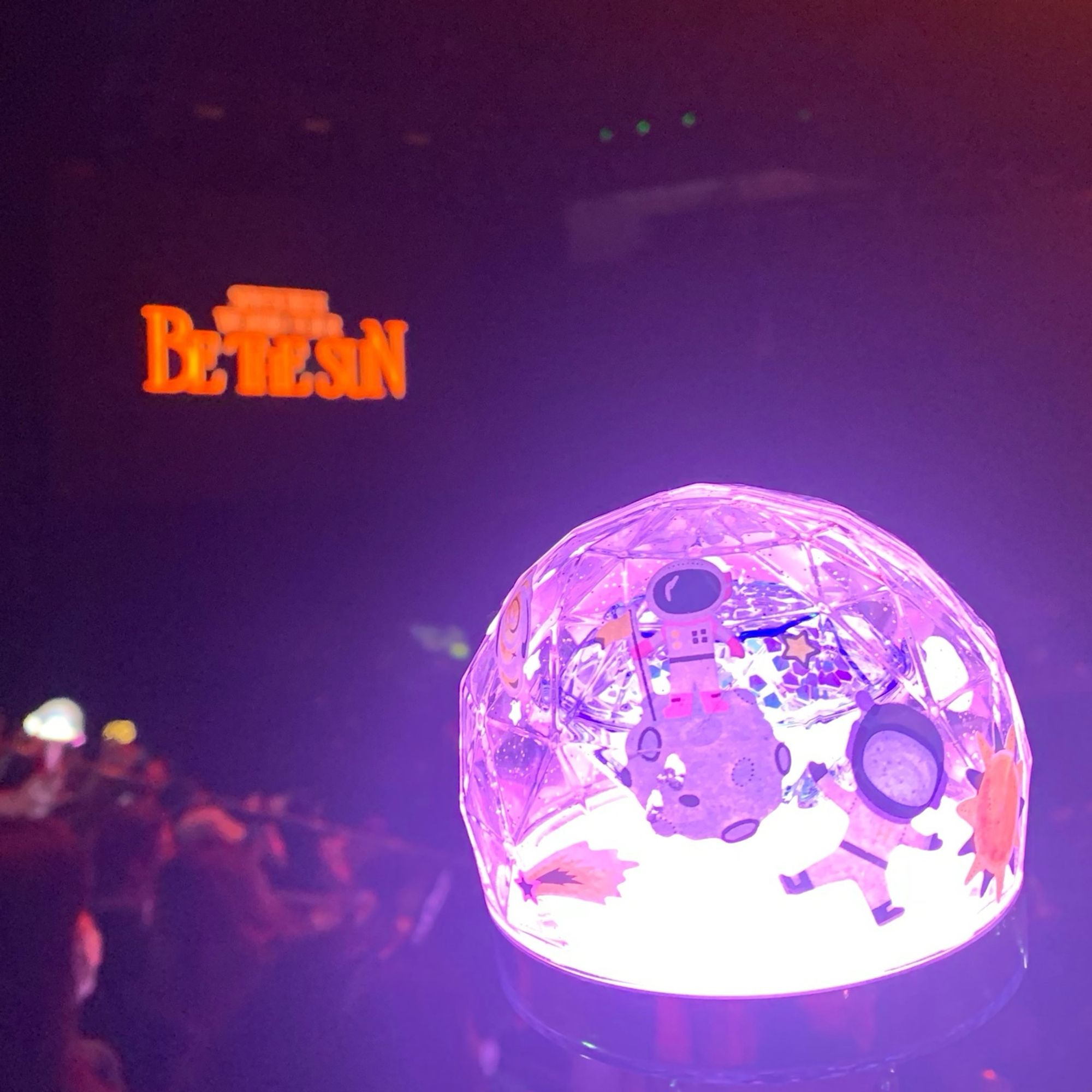 Photo of a Seventeen lightstick lit up in purple and decorated with astronaut stickers. An out of focus screen in the background reads “Be The Sun”