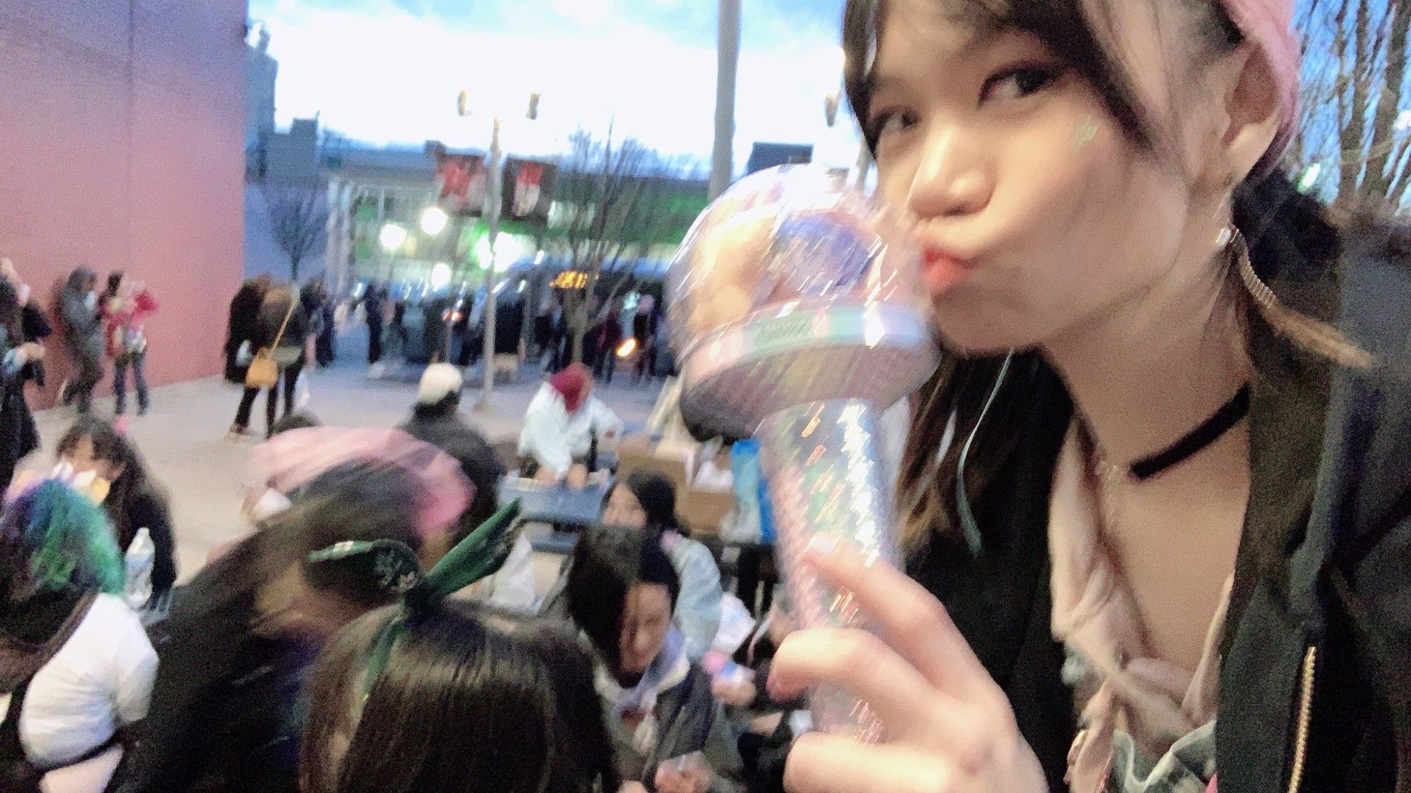 Photo of a dark haired person kissing the top of a Seventeen lightstick in a crowded outdoor space.