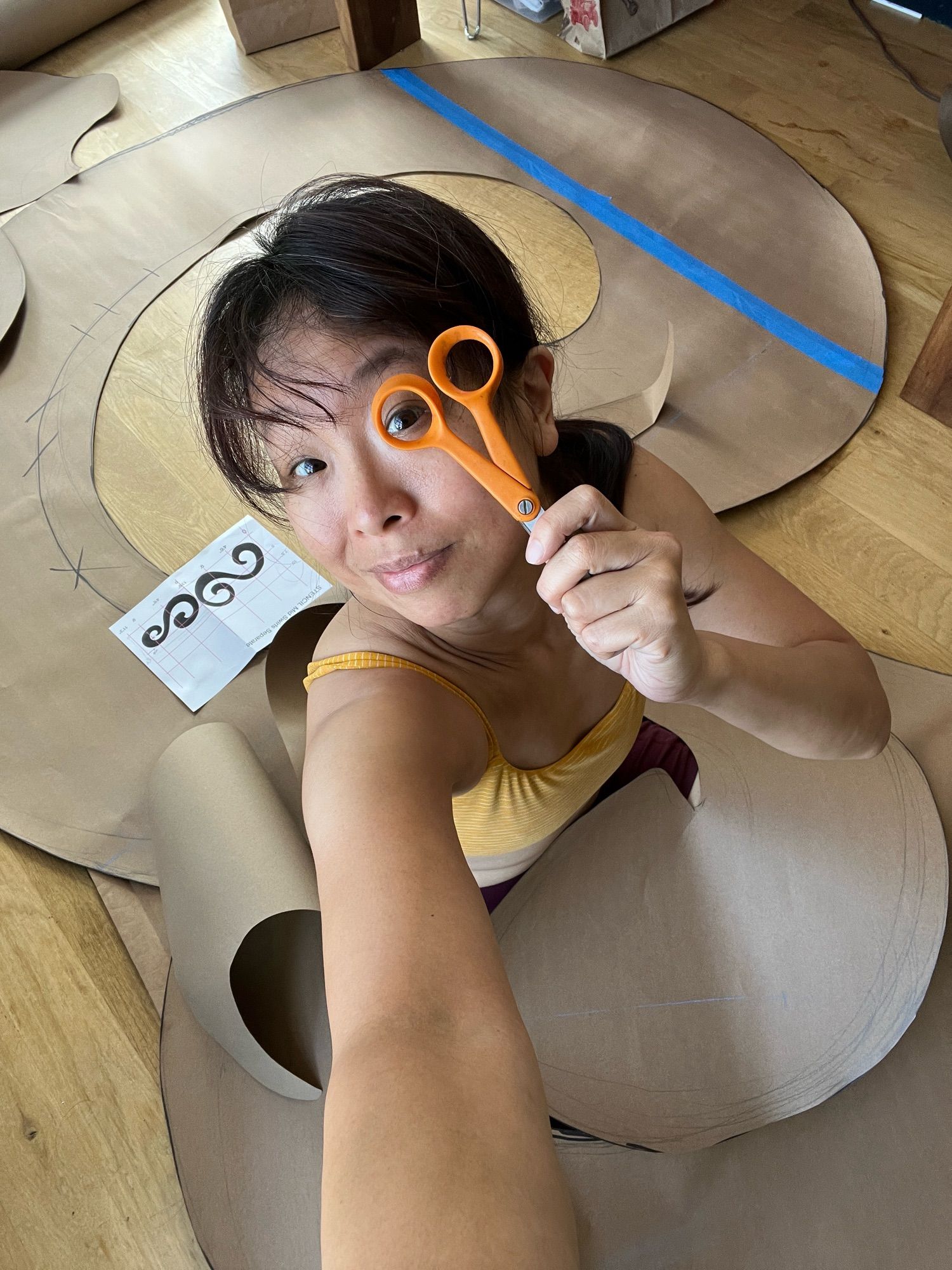 Artist looks through orange scissors as she sits on wooden floor with a giant swirl of Kraft paper piled around her