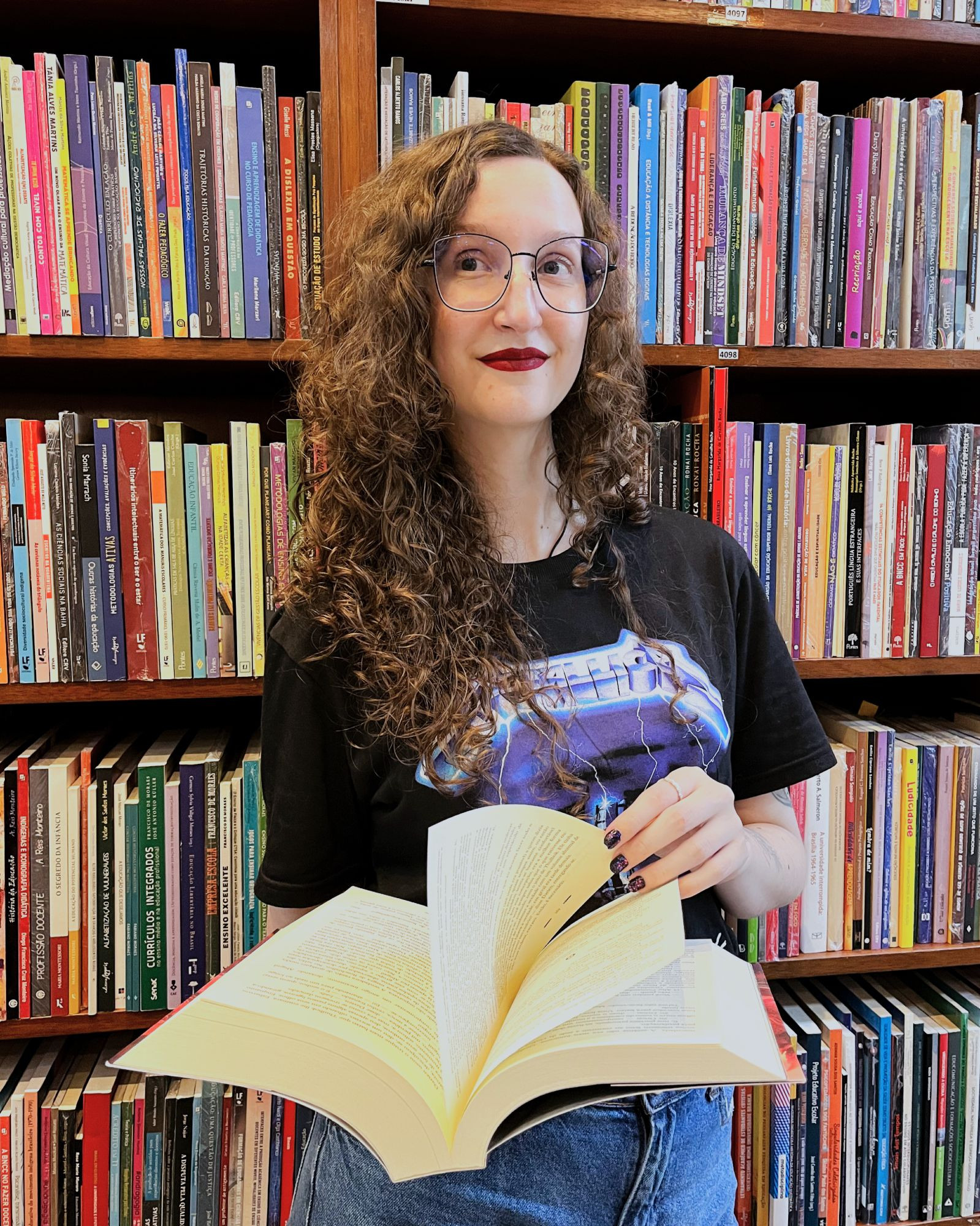 fotografia minha com uma estante de fundo. Estou segurando um livro com as páginas abertas. Tenho pele branca, cabelo cacheado loiro escuro e uso óculos de grau. Estou usando batom vermelho, sorrindo de leve e olhando para o lado. Estou usando uma camiseta preta do Metallica e calça jeans.