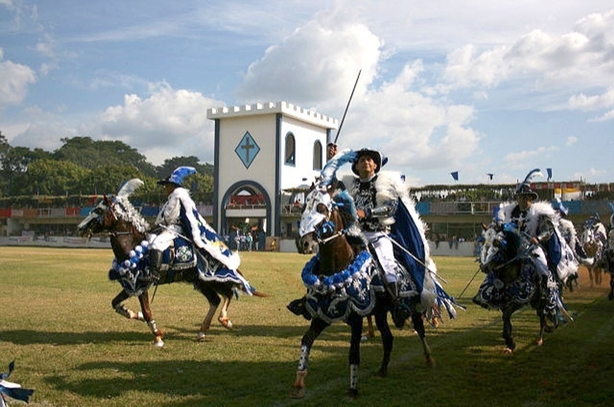 Fotografia colorida da apresentação das Cavalhadas de Pirenópolis (GO), com homens montados a cavalo enfeitados com roupas parecidas com as de mosqueteiros nas cores azul e branca, representando os cristãos na luta contra os Mouros.