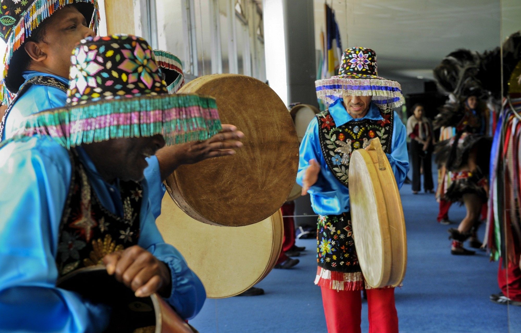 Fotografia colorida. Em espaço fechado, um grupo de bumba meu boi se apresenta. Em primeiro plano estão os percussionistas, segurando pandeirões na vertical e usabdo roupas e chapéis enfeitados de canutilhos coloridos que formam desenhos de flores folhas e pássaros. Ao fundo dançam outros membros do grupo, também enfeitados com penas ou chapéus bem maiores, com fitas longas que se estendem quase até o chão.