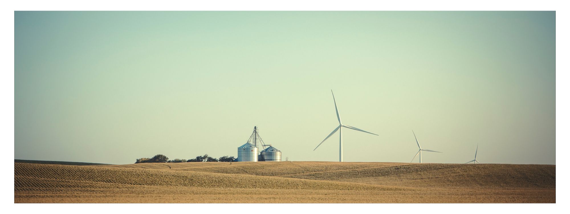 wide shot of golden tan fields in the late afternoon sun, with a distant silver shining grain silo & 3 wind turbines receding into the background. Pale blue sky - calm image