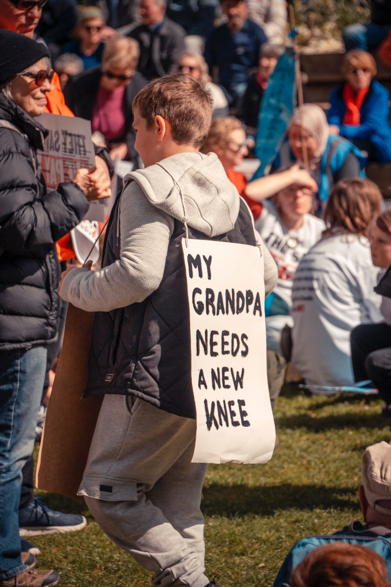 A kid wearing a sandwich board with the words 'My Grandpa needs a new knee'