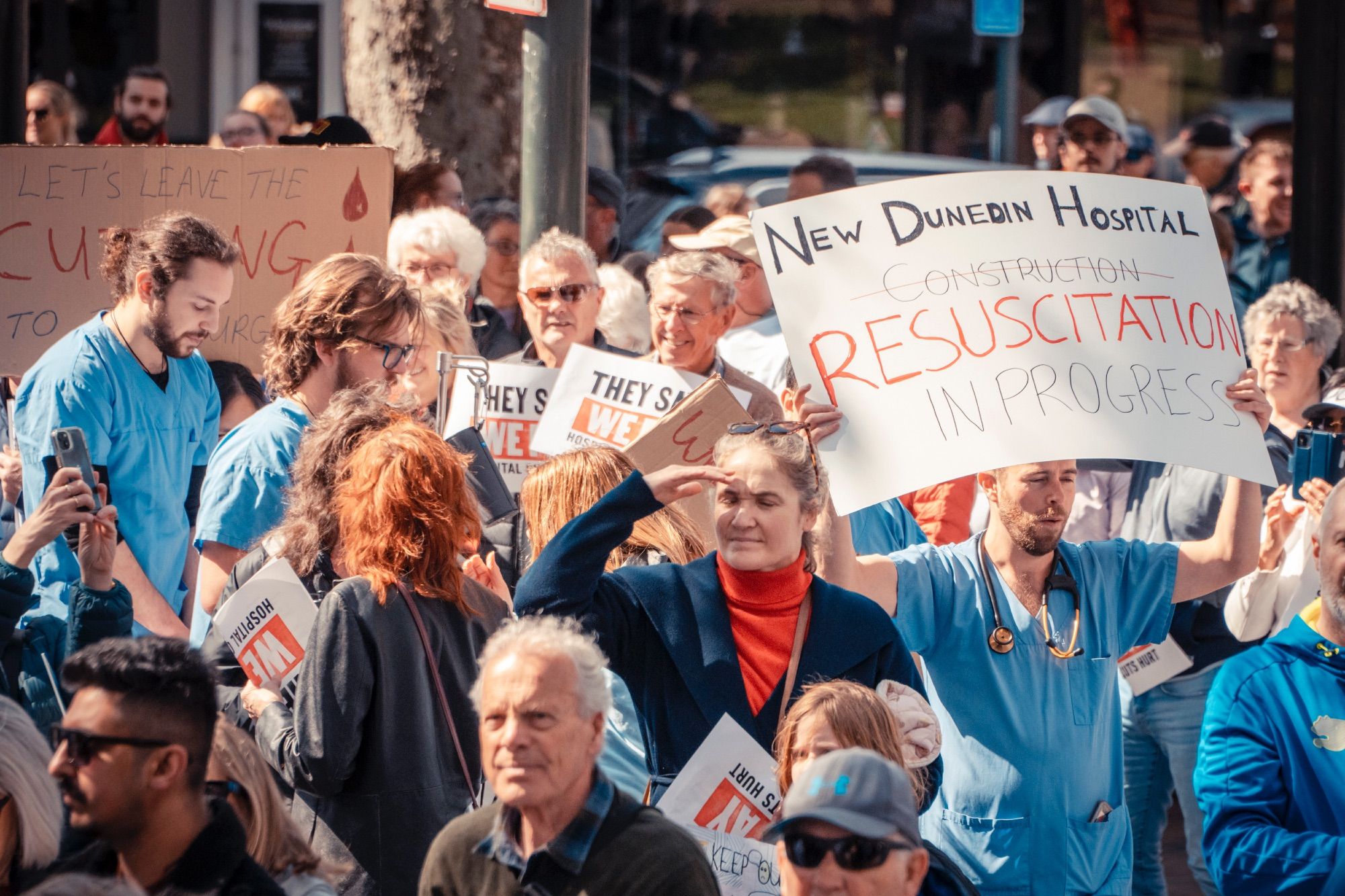 Med students in their scrubs holding a sign reading 

'New Dunedin Hospital
Construction [crossed out]
RESUSCITATION
IN PROGRESS'