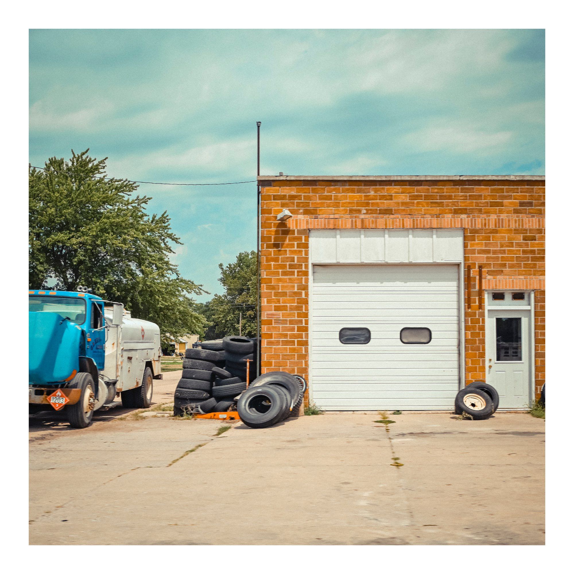 Small town USA garage store.  Broken down blue truck on left, pile of tyres, brick building with closed roller door. Warm blue sky, orange bricks & over all still calm image