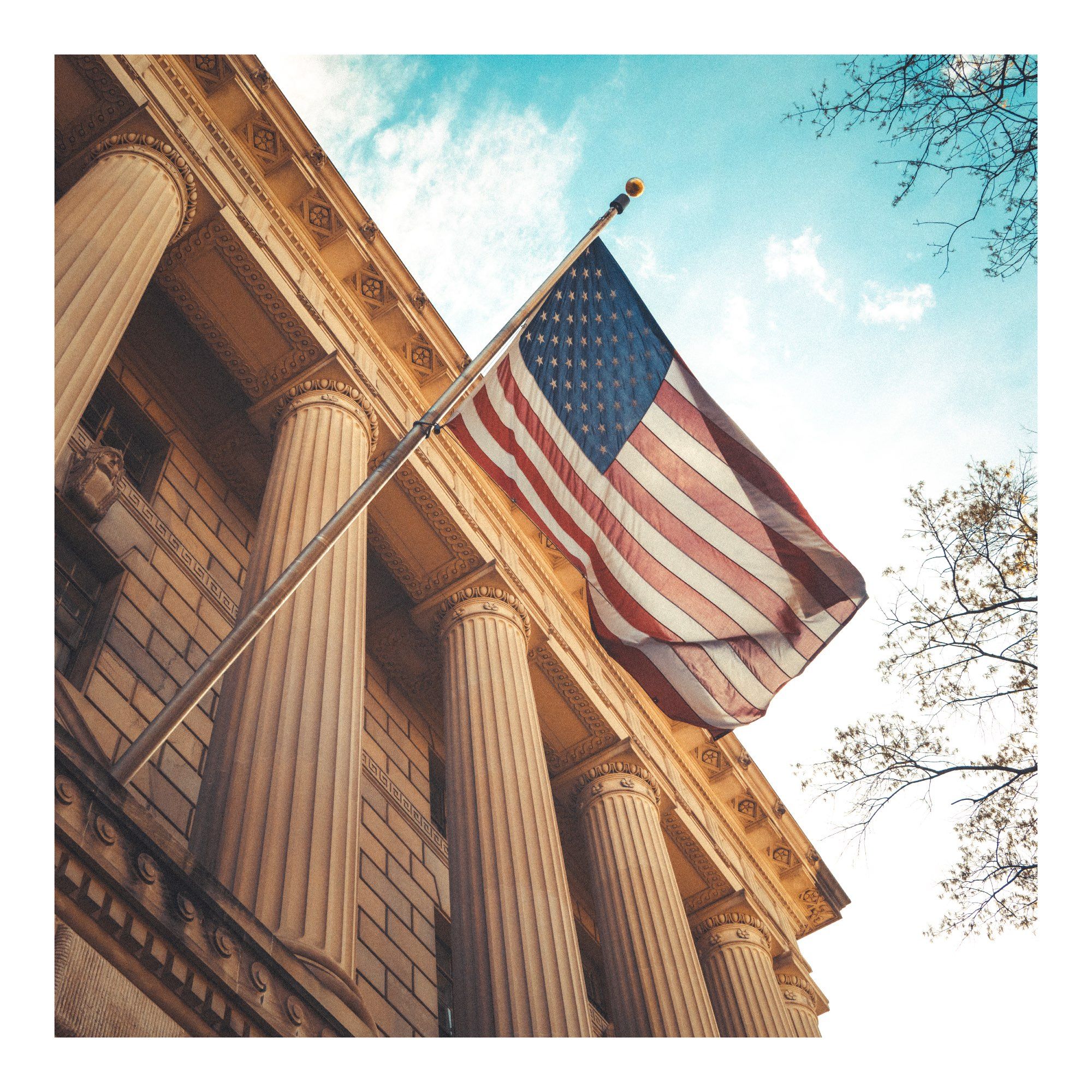 American flag outside a golden US federal building.  The building has magnificent doric columns.  Warm but muted tones