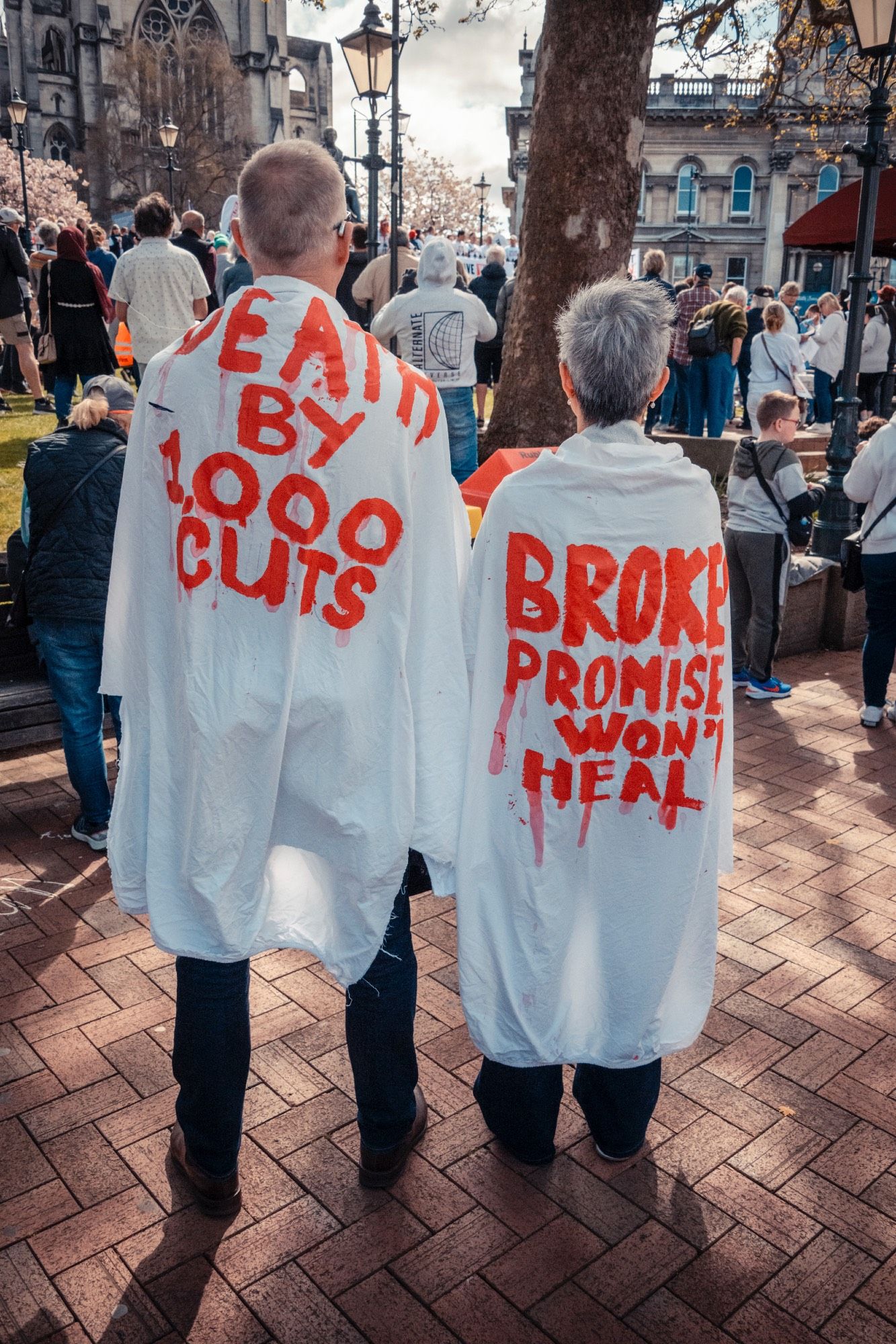 2 people wearing caps with words written in fake blood reading 

'Death by 1000 cuts' 
'Broken promises won't heal'