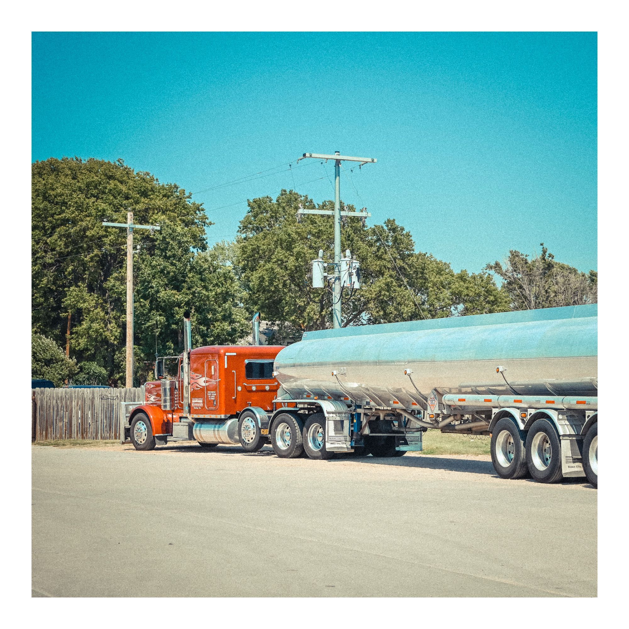 Classic American big rig tanker truck, with bright orange red double sleeper cab, long silver blue tank. Blue skies, pale green trees.  I think it's parked up for the evening.  Recreating pushing Ektachrome 100 film to 3200 so lots of grain.