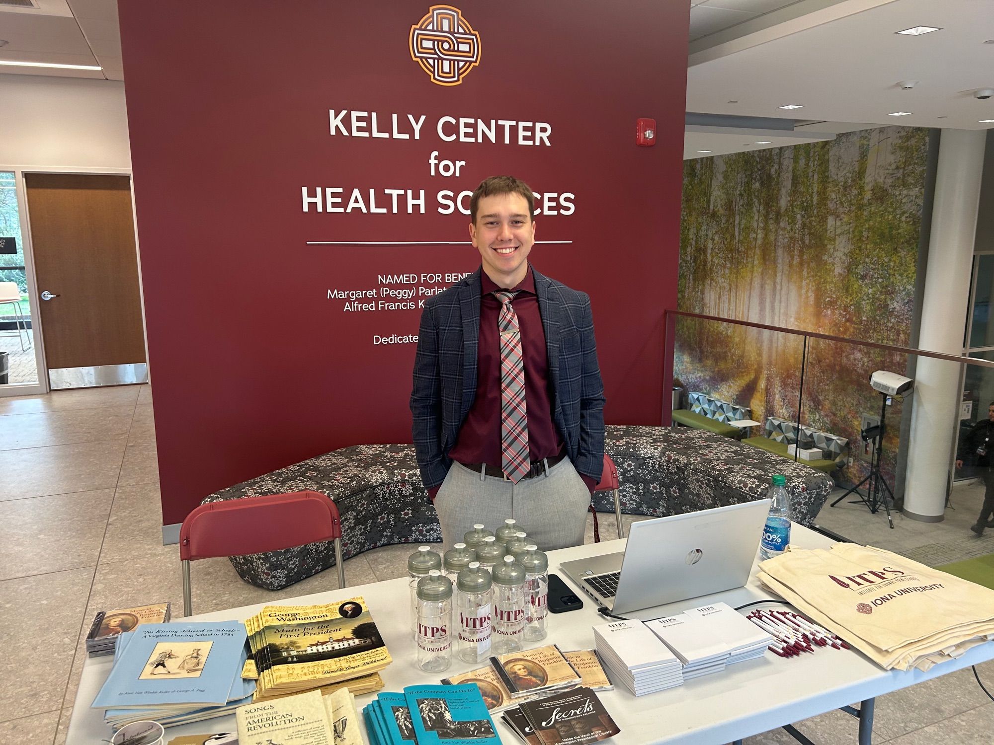 A student in a tie and blazer in front of a burgundy sign and folding table with giveaway items