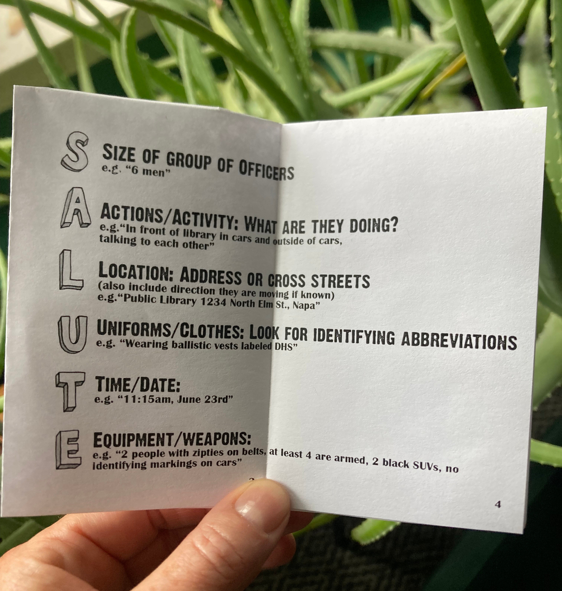 hand holding open zine. Pages read: "S Size of group, A Actions/Activity, L Location, U Uniforms/Clothes, T Time/Date, E Equipment