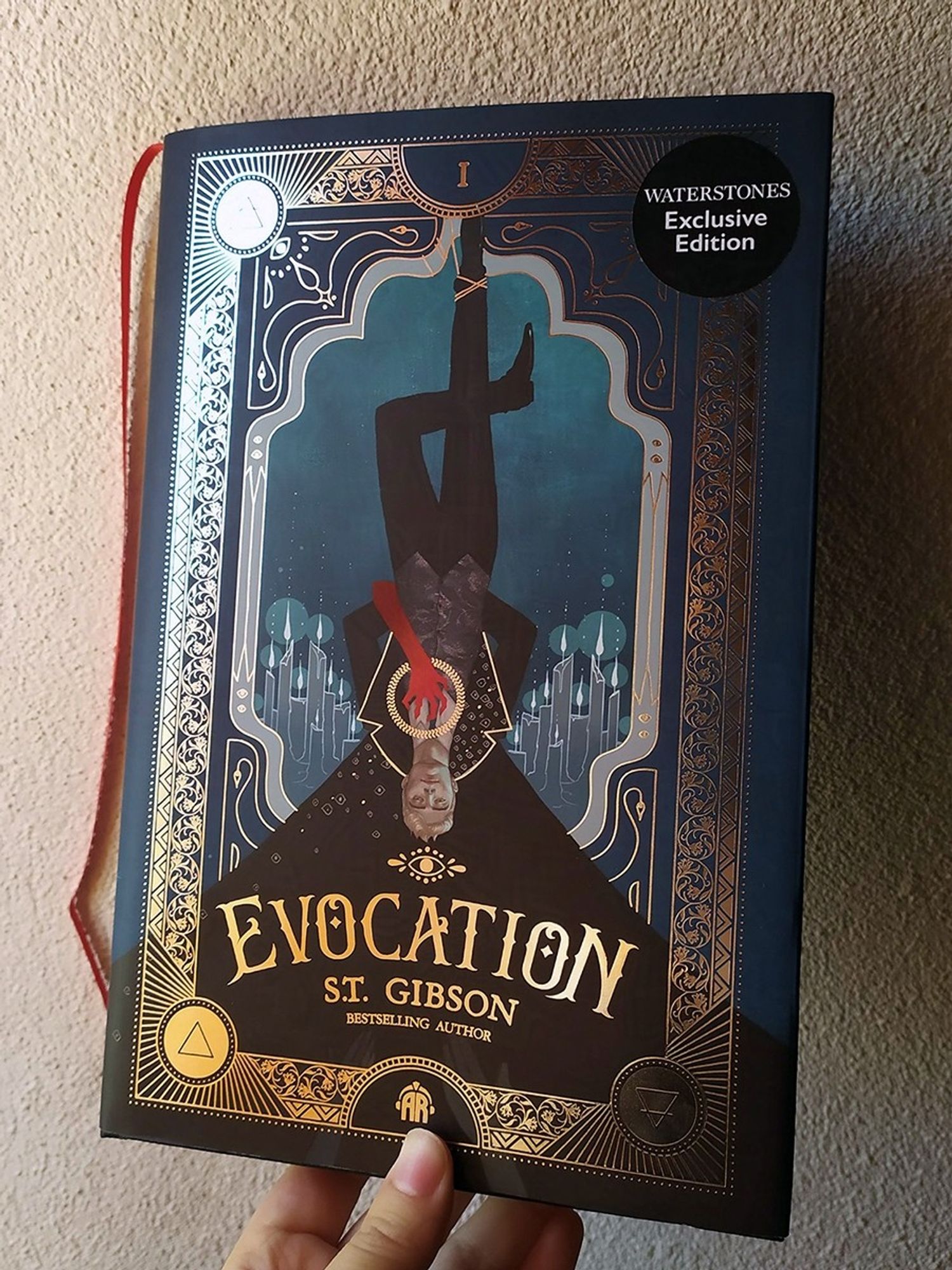 Photo of the book cover of EVOCATION by S.T. Gibson, featuring a man upside down with a mysterious red hand grabbing his chest. Illustration by Eleonor Piteira, design by Alice Claire Coleman.